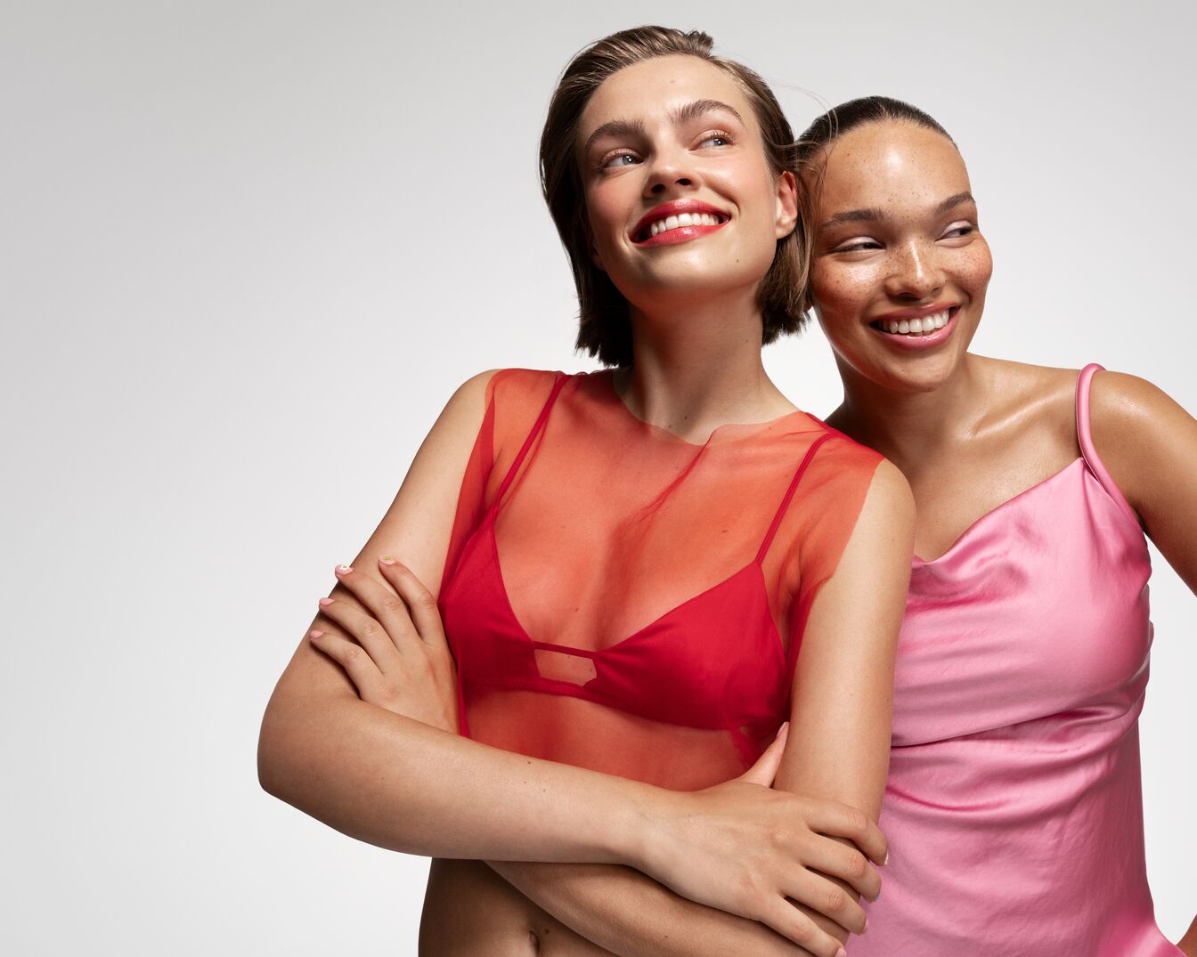 Zwei strahlende Frauen, eine in einem roten Oberteil und die andere in einem rosa Seidenkleid, blicken lächelnd zur Seite.