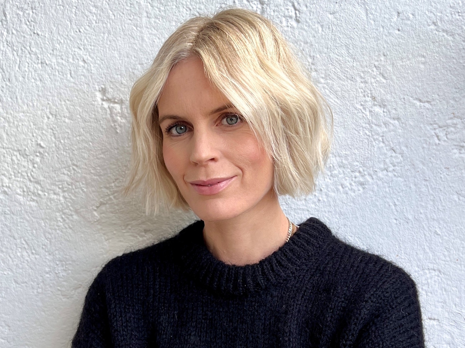 Porträt einer Frau mit blondem, gewelltem Bob und blauen Augen, die einen schwarzen Pullover vor einem weißen Hintergrund trägt.