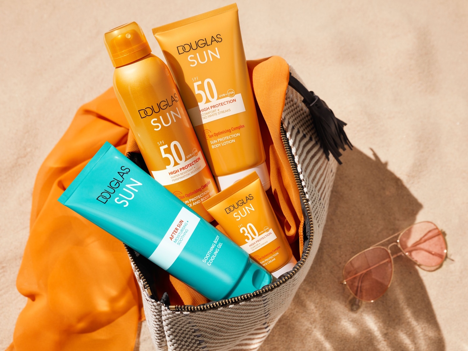 Verschiedene Sonnenschutzprodukte von Douglas Sun, darunter Sprays, Lotionen und Gels mit LSF 30 und 50, in einer Strandtasche mit einer orangefarbenen Jacke und einer Sonnenbrille.
