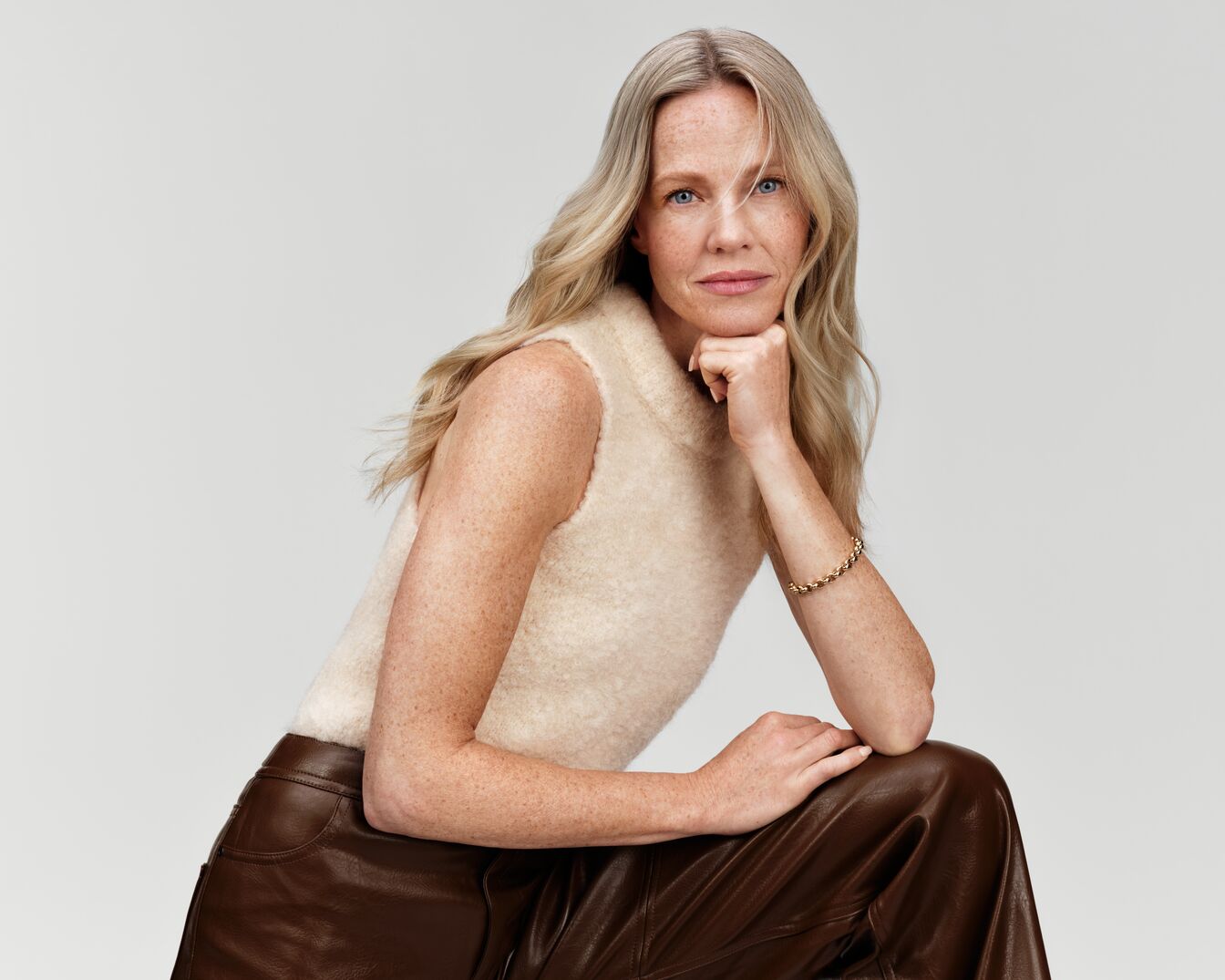 Eine Frau mit blonden, gewellten Haaren, einem ärmellosen Oberteil und einer braunen Lederhose posiert mit einem goldenen Armband.