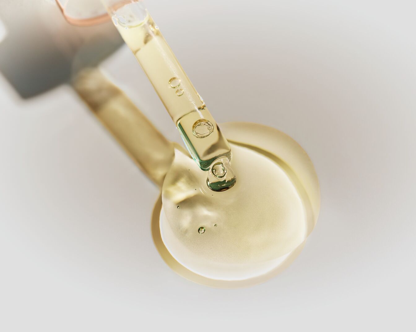 Nahaufnahme eines durchsichtigen Glaspipette, die eine goldfarbene Flüssigkeit auf eine glatte, goldene Oberfläche tropft.