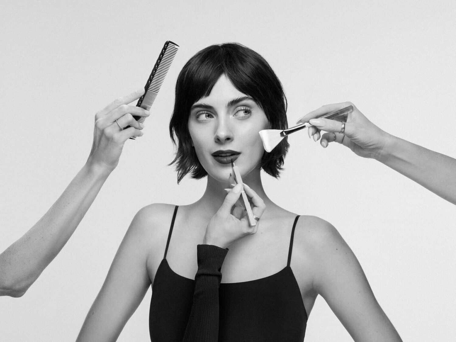 Juodai balta nuotrauka, kurioje moteris su juoda suknele, kuriai daromas makiažas ir šukuosena, naudojant šepetėlį, pieštuką ir šukas.