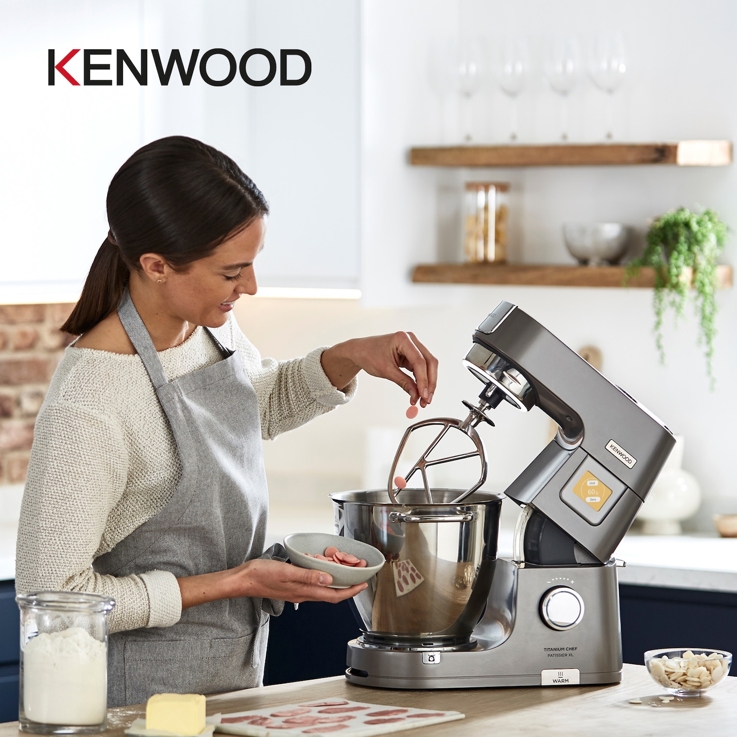 Eine Frau mit grauer Schürze fügt einem Kenwood Titanium Chef Patissier XL Mixer rosa Süßigkeiten hinzu, während Zutaten wie Mehl und Mandeln bereitstehen.