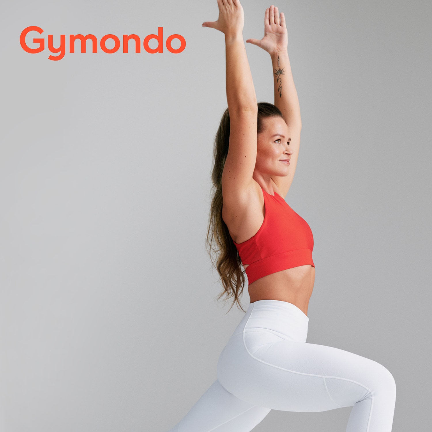 Eine Frau in Sportkleidung, rotes Top und weiße Leggings, macht Yoga vor einem grauen Hintergrund, mit dem orangefarbenen "Gymondo"-Logo oben links.