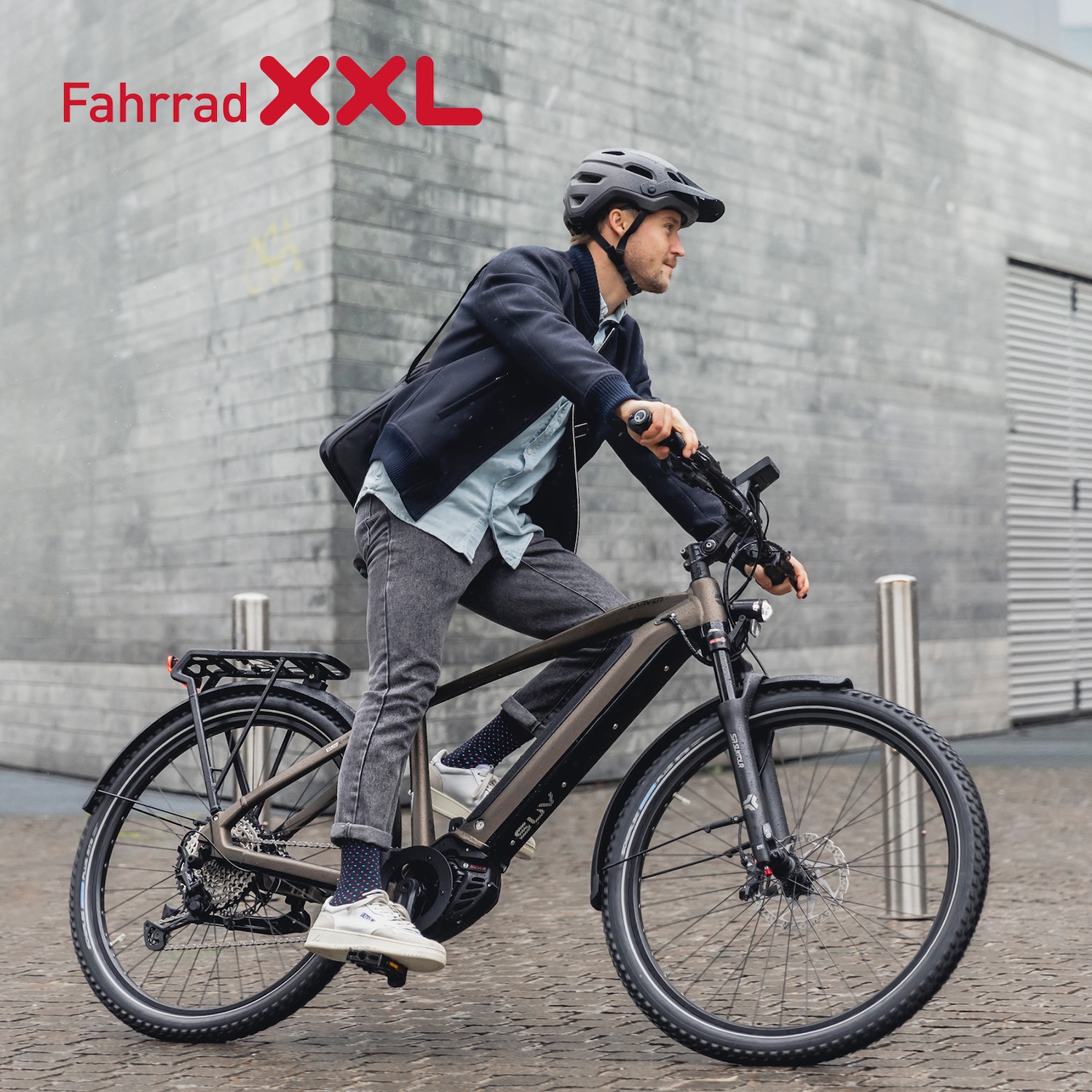 Ein Mann mit Helm fährt ein modernes, braunes E-Bike vor einer grauen Backsteinmauer, beworben mit dem roten Schriftzug Fahrrad XXL.