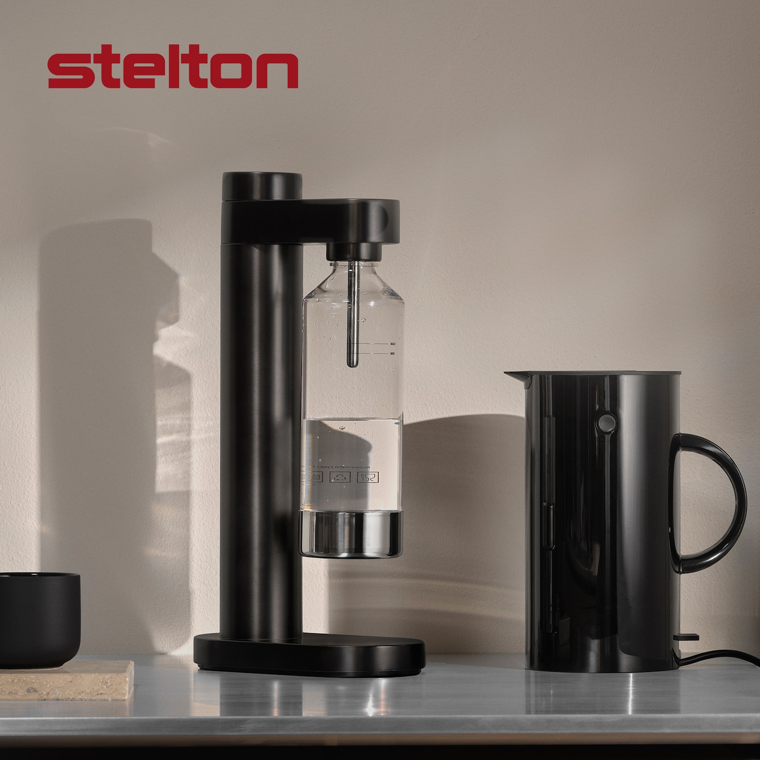 Stelton Wassersprudler in Schwarz mit Wasserflasche und schwarzem Krug auf einer Arbeitsfläche.