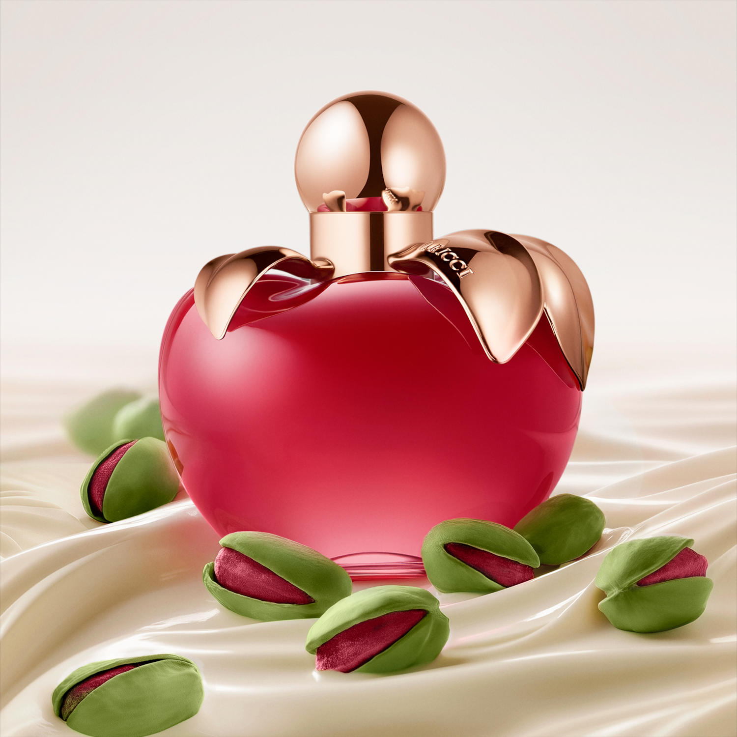 Découvrez le parfum Nina Ricci dans son flacon rouge en forme de pomme, orné de feuilles dorées, entouré de pistaches sur un tissu soyeux.