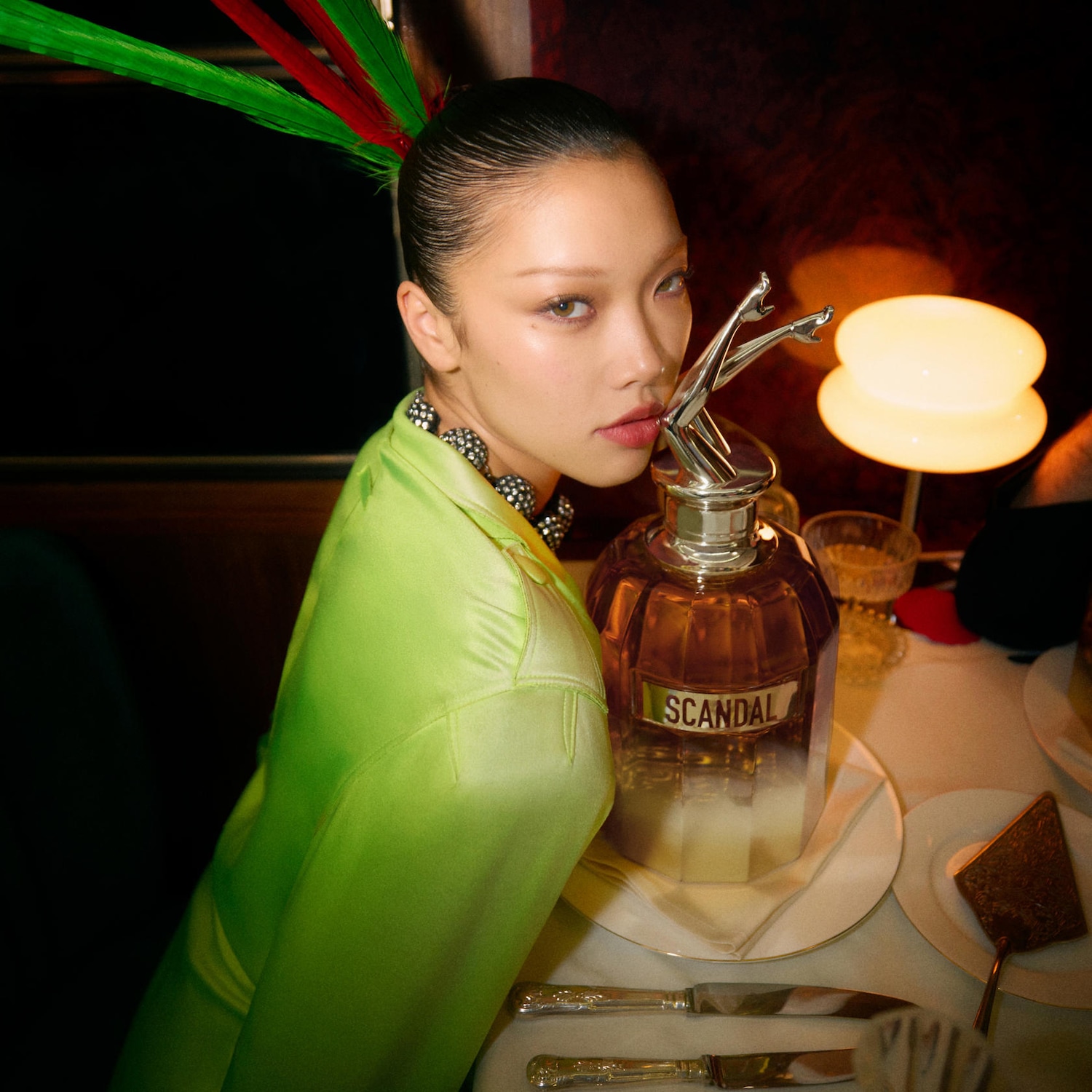 Une femme élégante en veste vert vif pose avec un flacon de parfum Scandal, coiffée de plumes colorées, créant une scène sophistiquée et audacieuse.