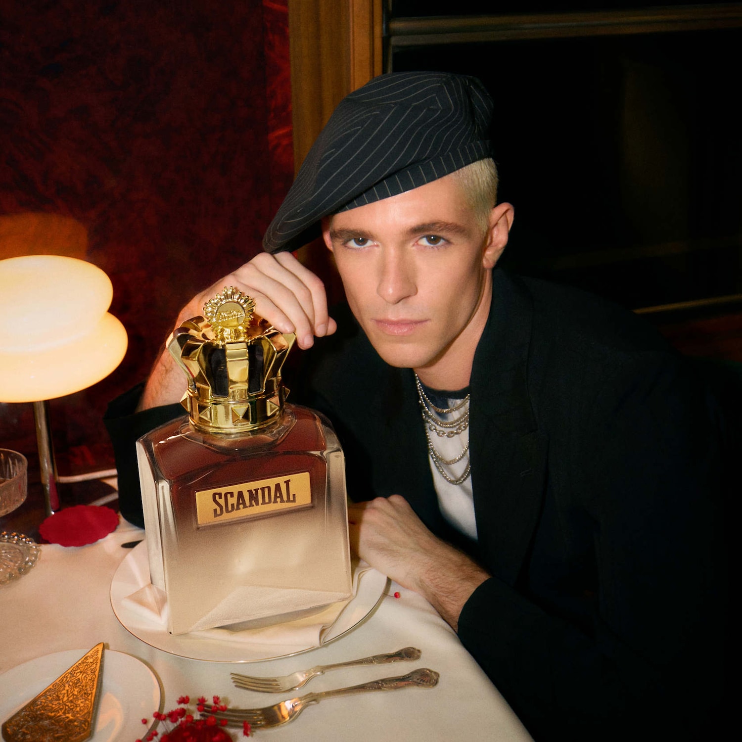 Un homme élégant pose avec le parfum Scandal, dont le flacon est surmonté d'une couronne dorée, évoquant un luxe audacieux et une sophistication moderne.