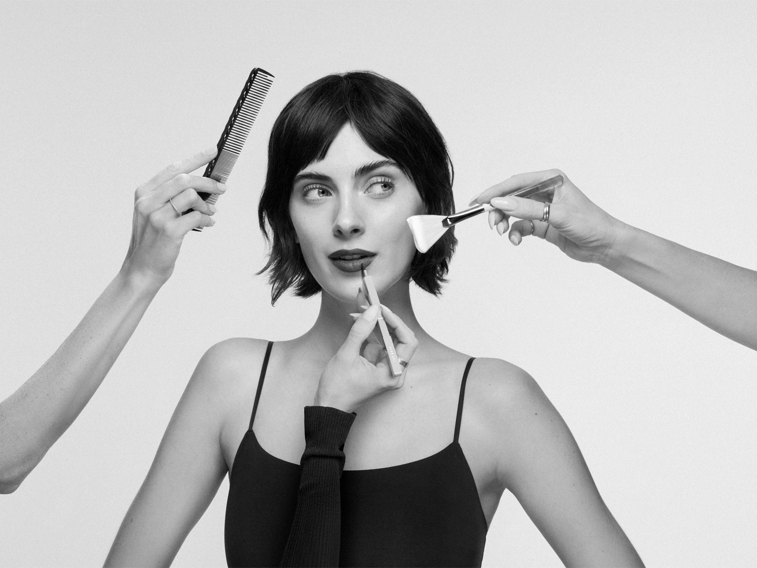 Primo piano in bianco e nero di una donna che applica il rossetto con un pennello, mentre una mano le sistema i capelli con un pettine.