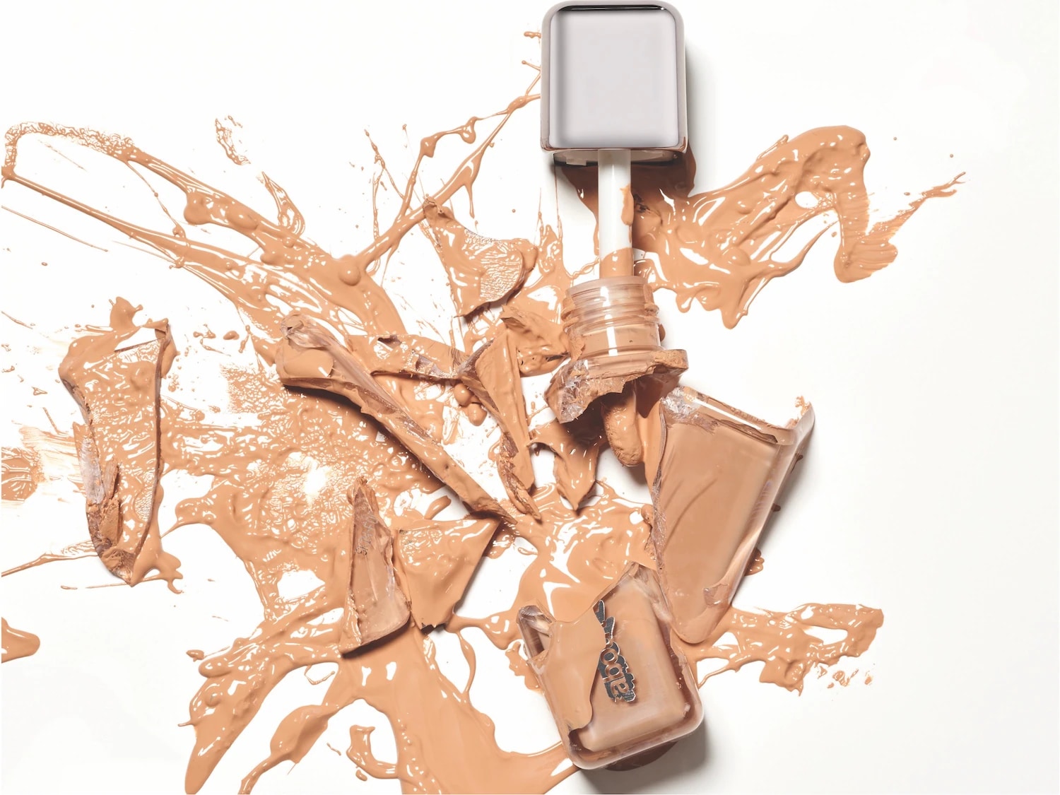 Een gebroken fles foundation met een beige kleur is te zien, de inhoud is uitgespat op een witte achtergrond.