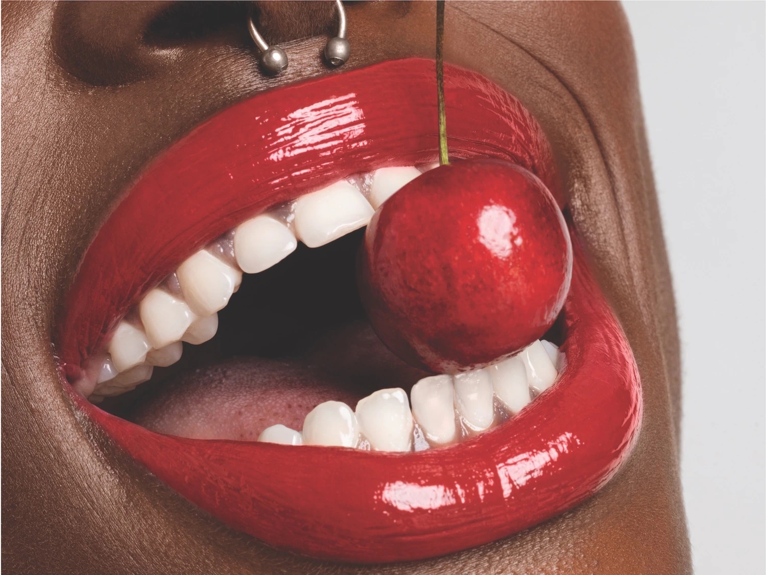 Een close-up van een vrouw met een neuspiercing, felrode lippenstift en een kers tussen haar tanden.