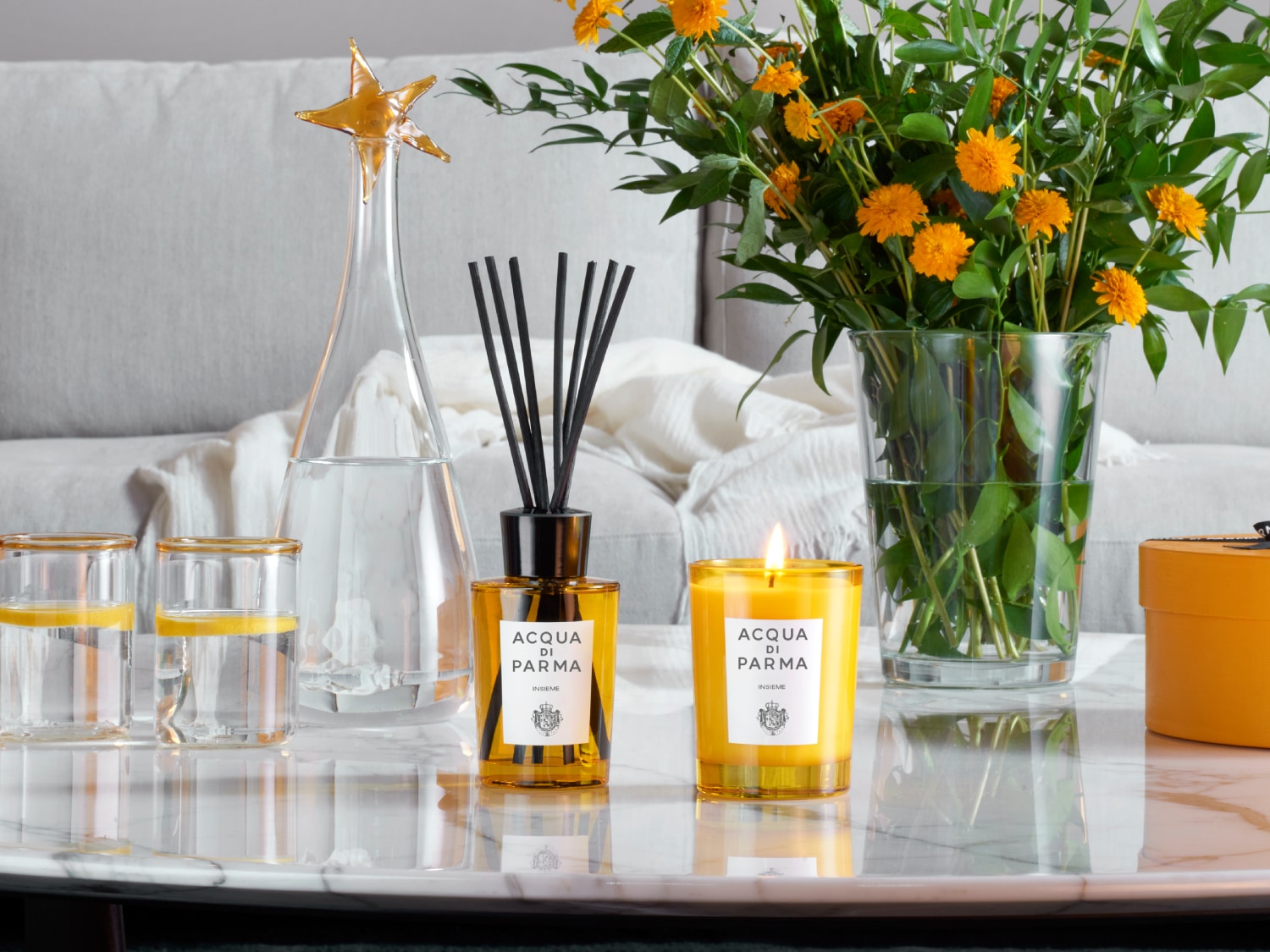 Einrichtung mit Acqua di Parma Duftkerze, Duftstäbchen und gelben Blumen auf einem Marmortisch, die ein gemütliches Ambiente schaffen.
