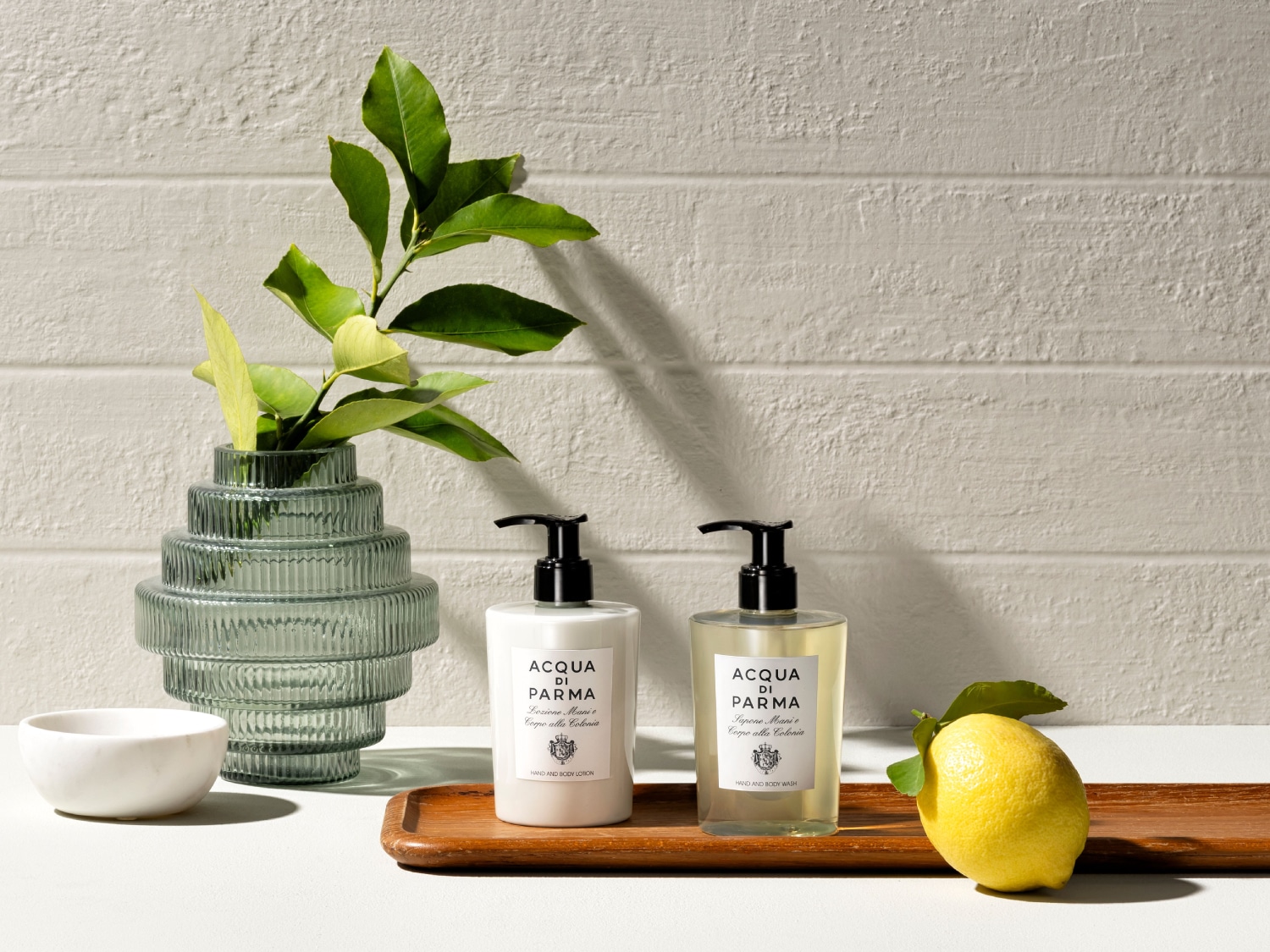 Auf einem Tisch stehen eine Vase mit grünen Blättern, eine weiße Schale, eine Handlotion und eine Handwaschseife von Acqua di Parma sowie eine Zitrone.