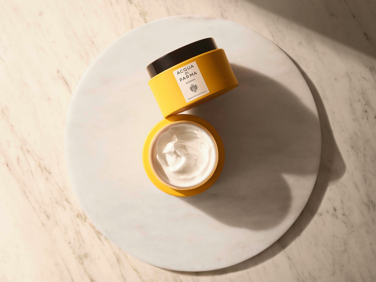 Dargestellt sind zwei gelbe Tiegel der Marke Acqua di Parma, einer davon geöffnet, um die weiße Creme im Inneren zu zeigen.