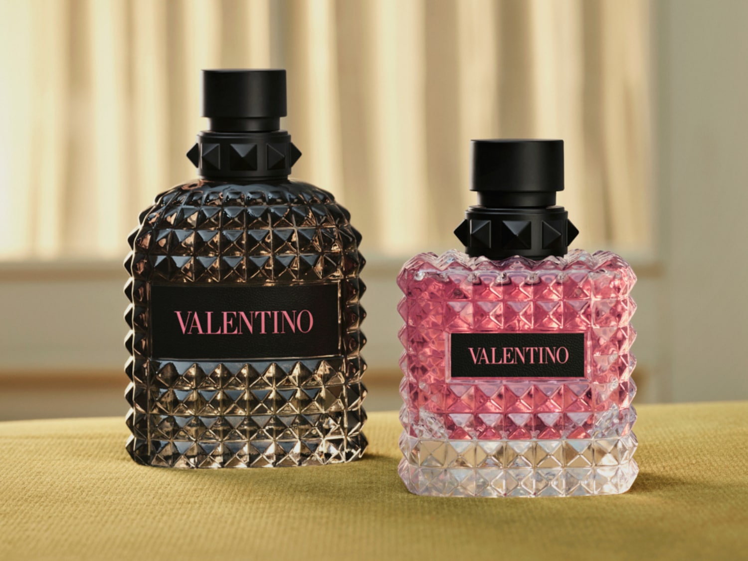 Két Valentino parfümös üveg látható, az egyik sötét, a másik rózsaszín, mindkettő fekete kupakkal és Valentino felirattal.
