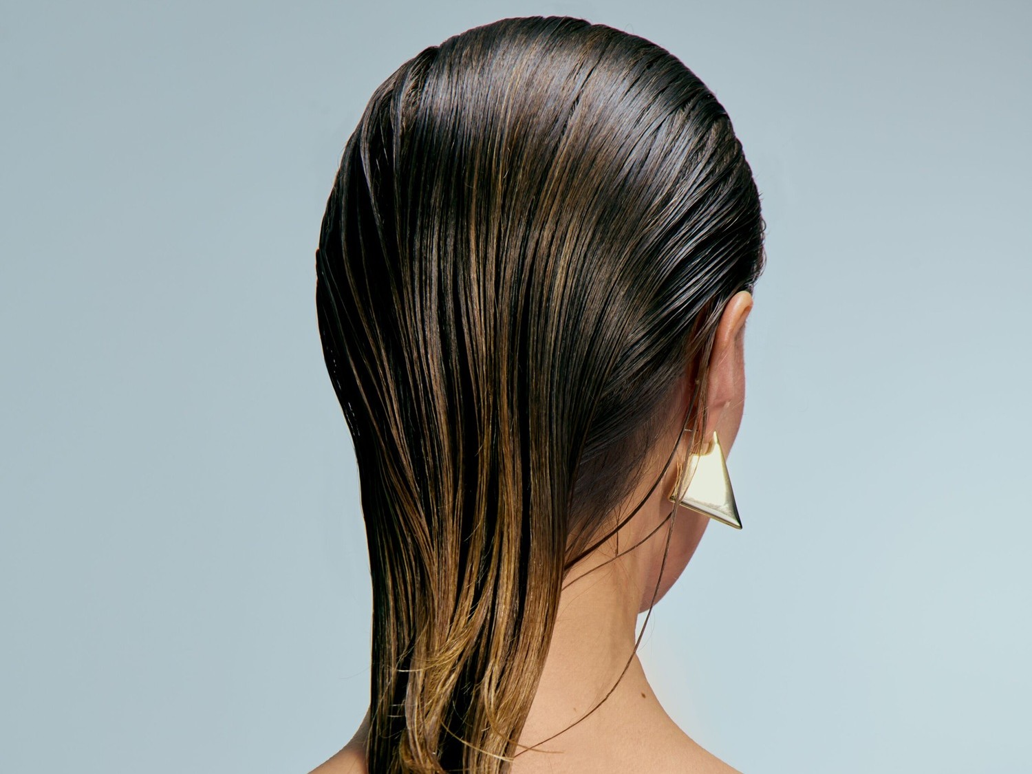 Nahaufnahme der Rückansicht einer Frau mit glänzendem, dunkelbraunem Haar, das zu den Spitzen hin heller wird, und einem goldenen, dreieckigen Ohrring.