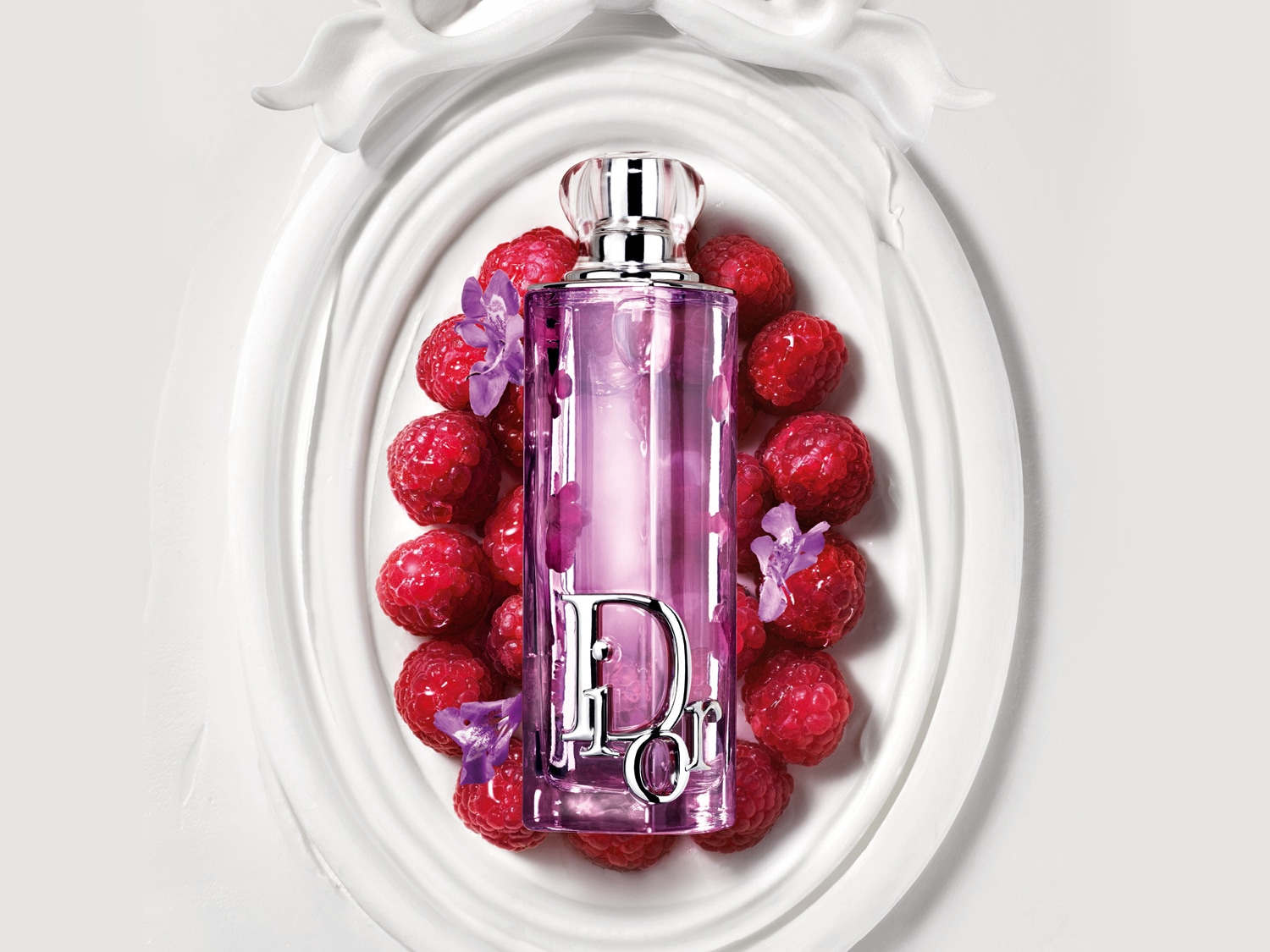 Su un piatto bianco decorativo, un flacone di profumo Dior rosa è circondato da lamponi rossi e fiori viola.