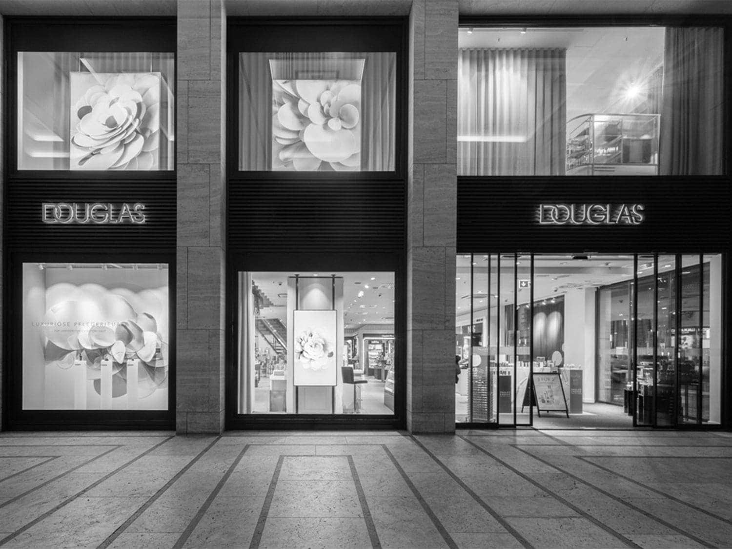 Fachada en blanco y negro de la tienda Douglas con grandes escaparates que exhiben flores decorativas y el logo luminoso de la marca.