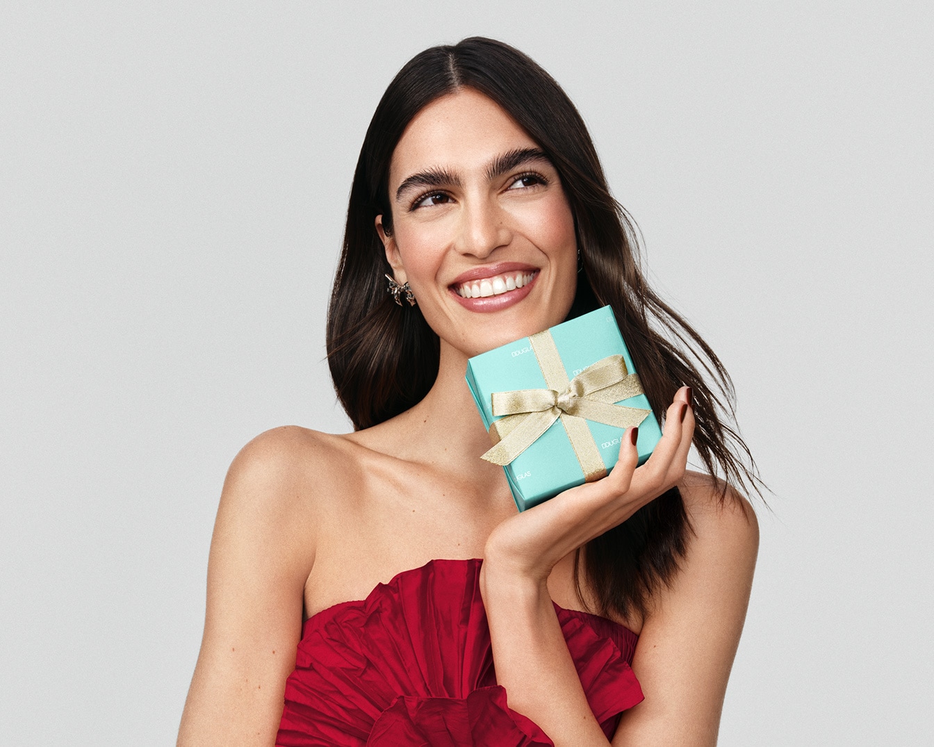 Eine strahlende Frau im roten Kleid hält eine hellblaue Geschenkbox mit goldener Schleife und lächelt erwartungsvoll.