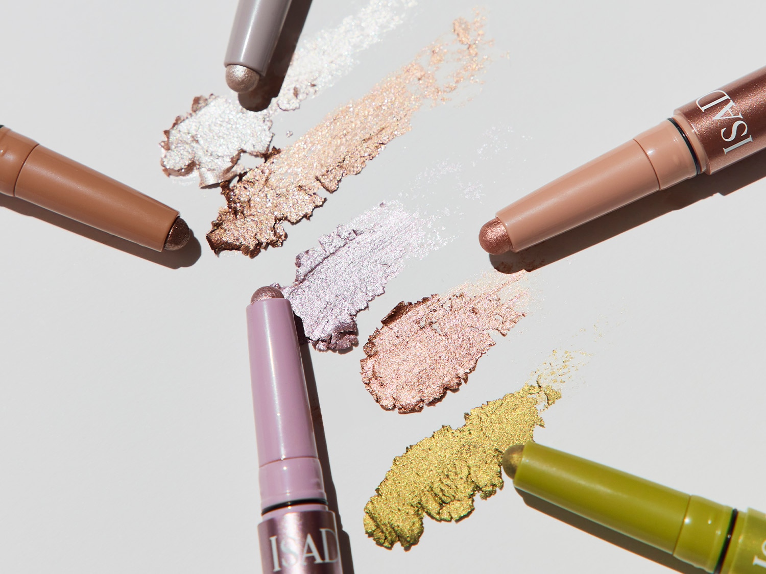 Aquí hay lápices de sombra de ojos ISAD en tonos marrón, rosa, morado, verde y gris, junto con muestras de los colores.