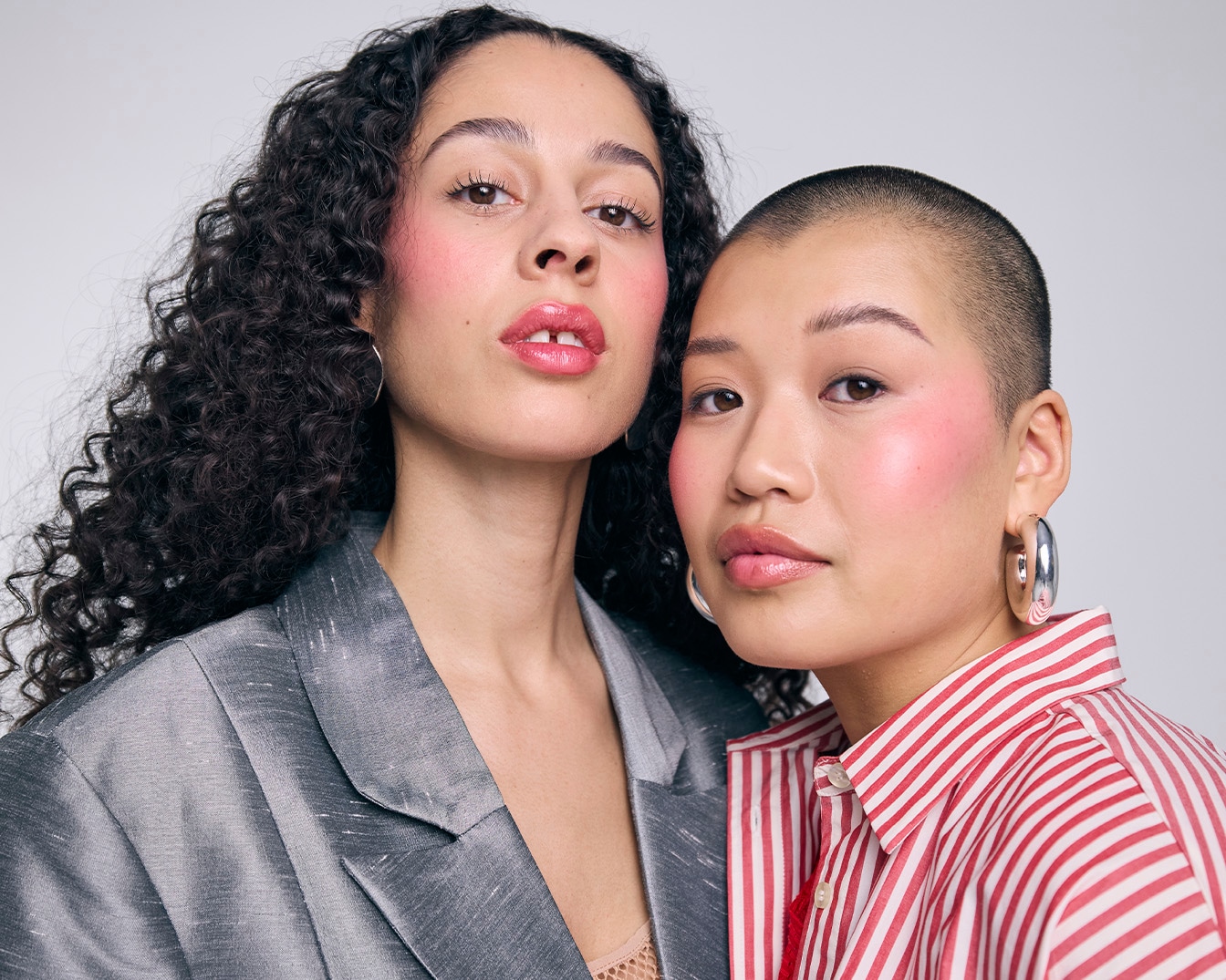 Dos modelos con maquillaje rosado, una con cabello rizado y chaqueta gris, la otra con cabello corto y camisa a rayas rojas y blancas, ambas con pendientes plateados.
