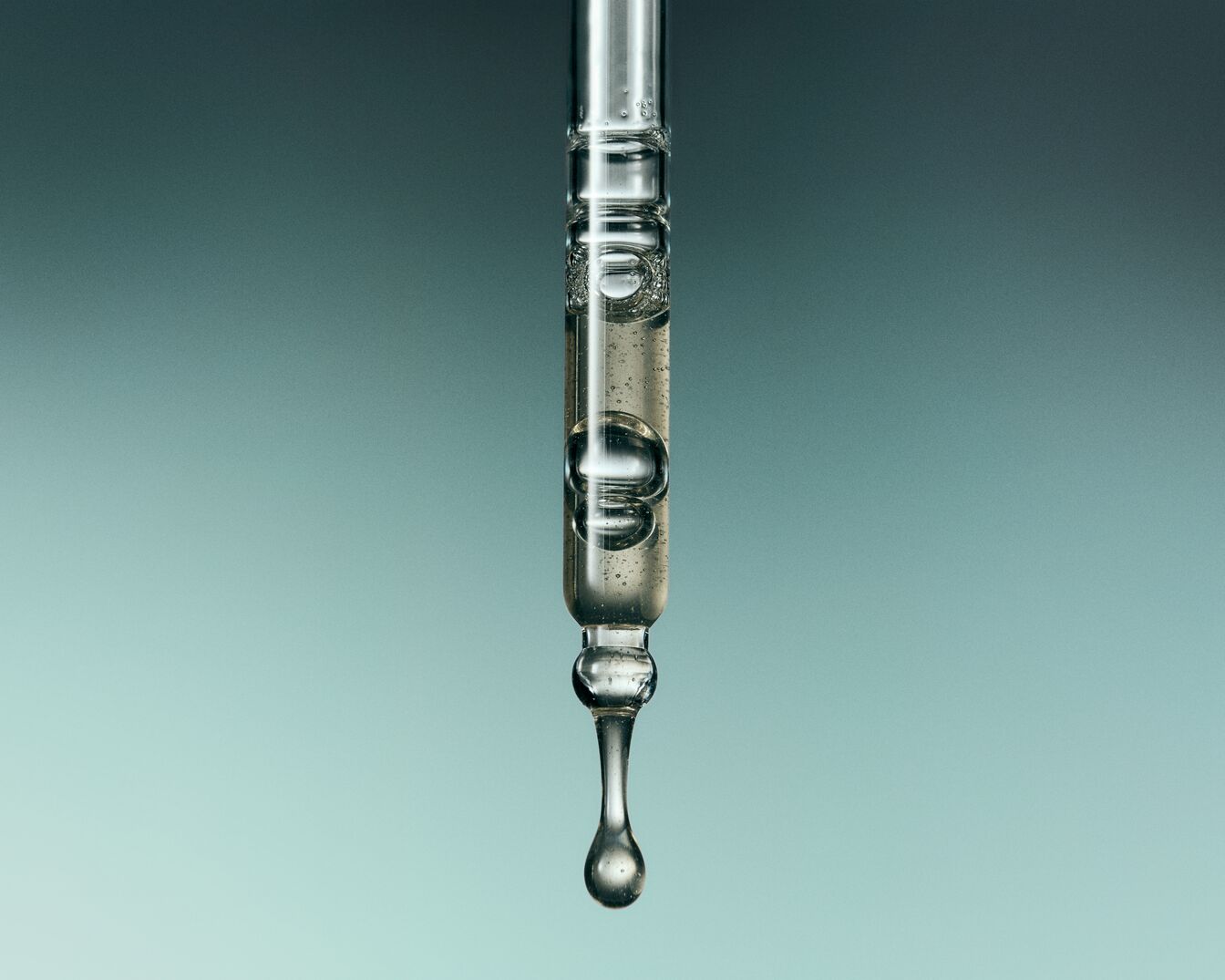 Primo piano di un contagocce di vetro trasparente pieno di siero liquido con bolle d'aria, che mostra una goccia che sta per cadere, su uno sfondo sfumato blu-verde.