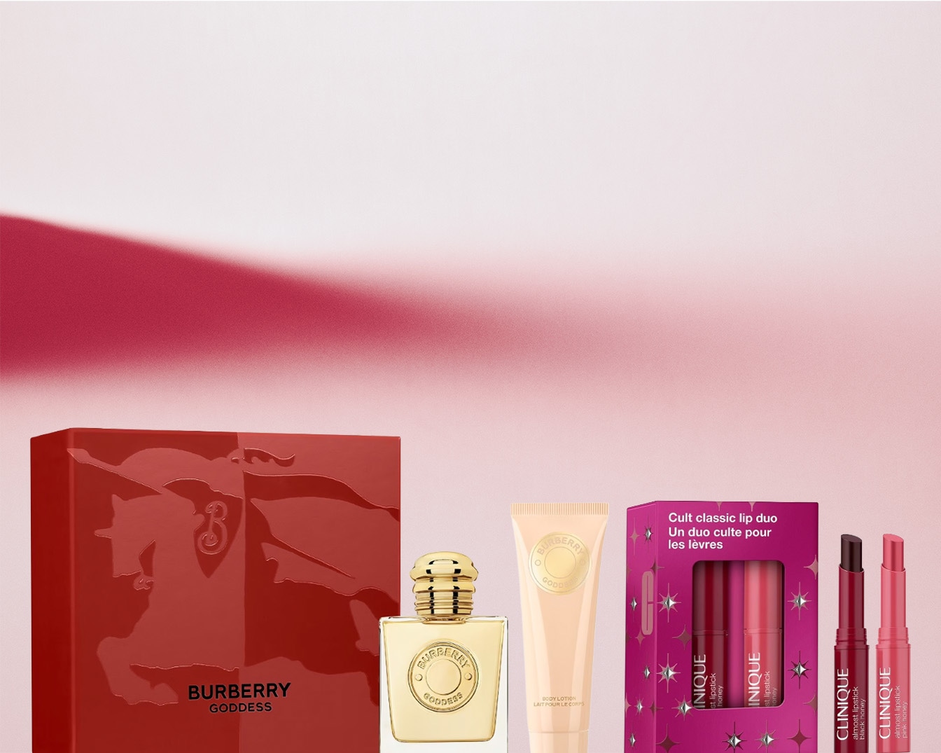 Confezione regalo Burberry Goddess color rosso vivo, affiancata da un flacone di profumo dorato, una lozione corpo e un duo di rossetti Clinique.