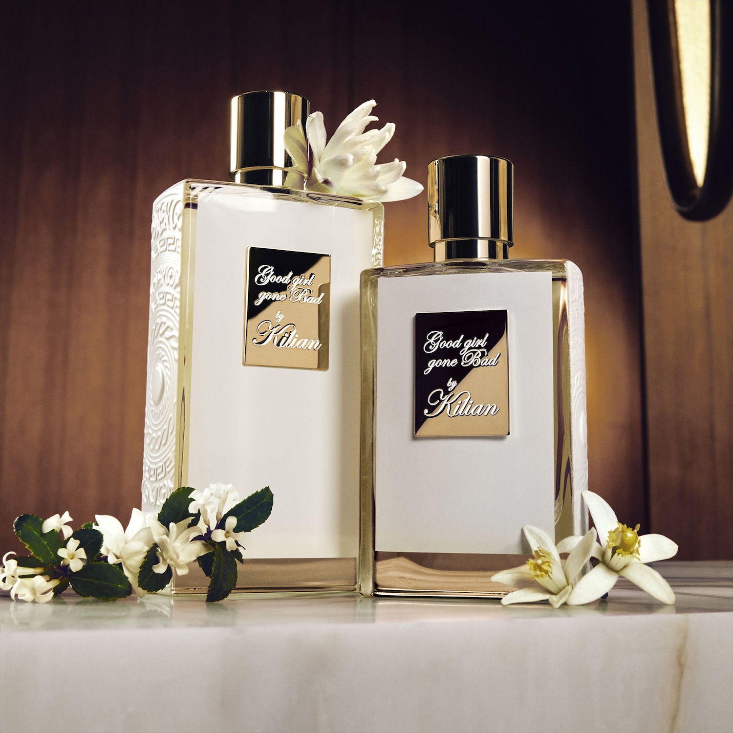 Twee flessen Kilian Good Girl Gone Bad parfum, de ene met een gegraveerde zijkant en de andere glad, beide met gouden accenten en witte bloemen.