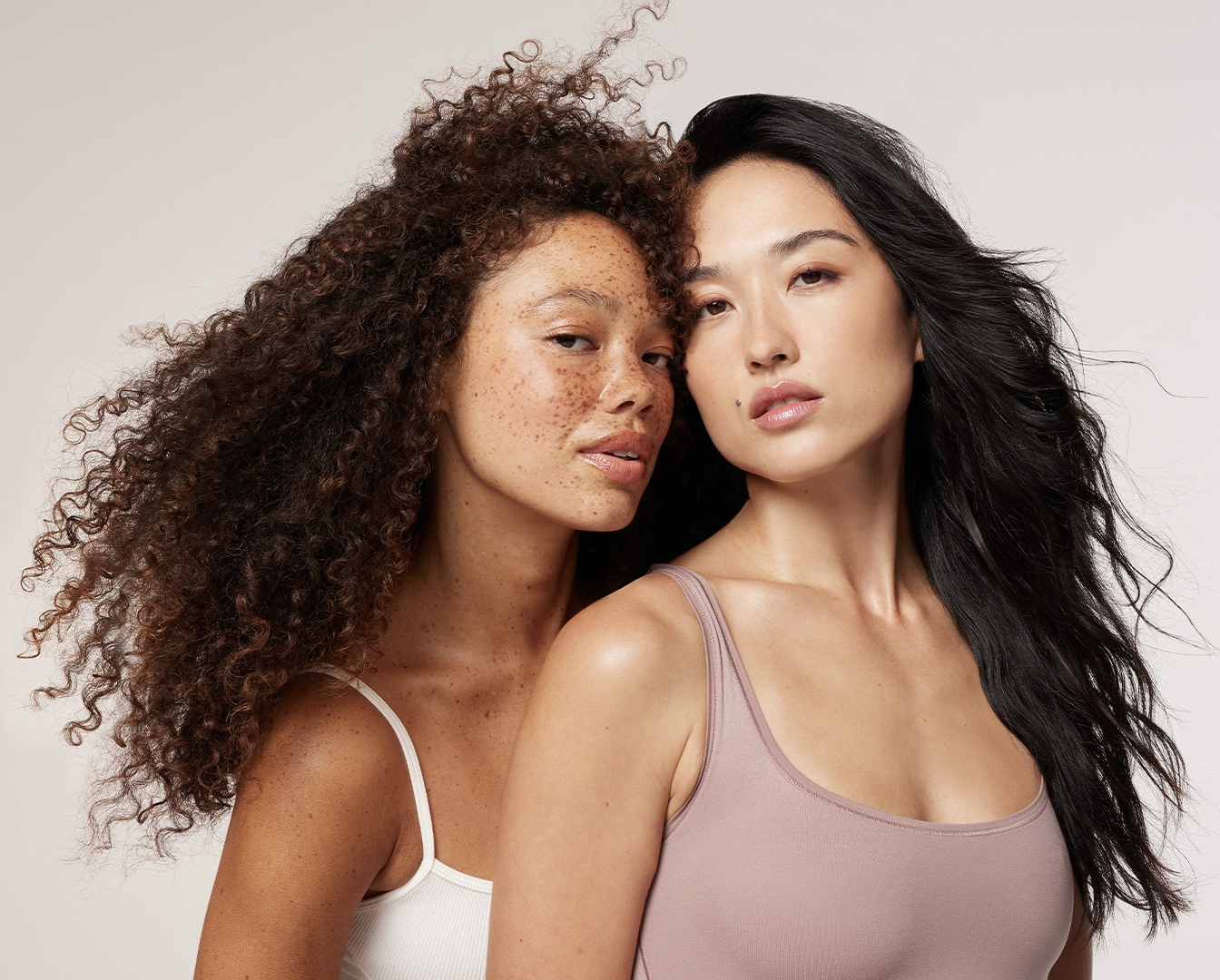 Twee vrouwen met verschillende huidtinten en haartexturen staan dicht bij elkaar, de een met krullend haar en sproeten, de ander met steil, donker haar.