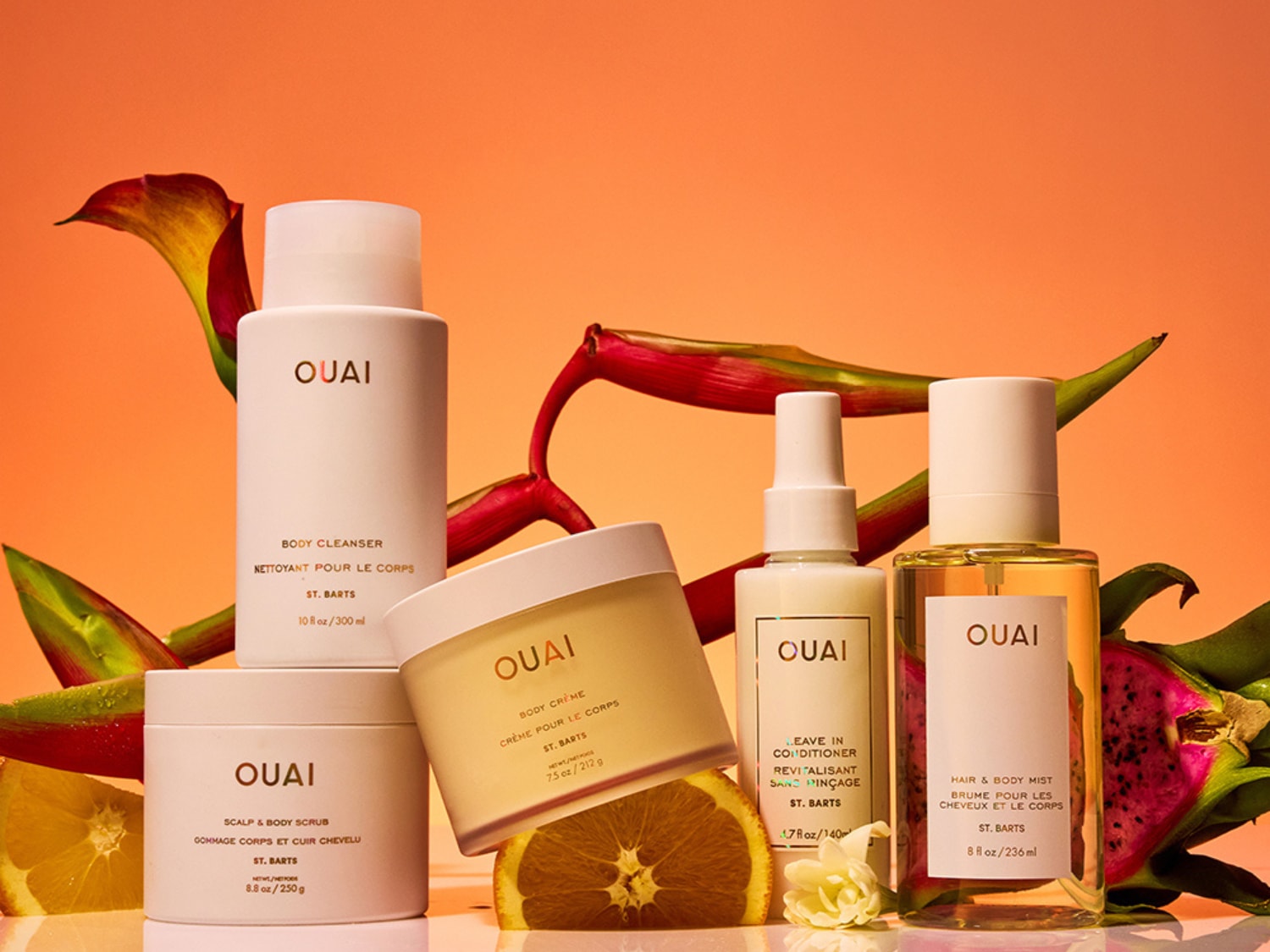 Ontdek de OUAI St. Barts collectie: body cleanser, scrub, crème, leave-in conditioner en hair & body mist, gepresenteerd met tropische bloemen en fruit.