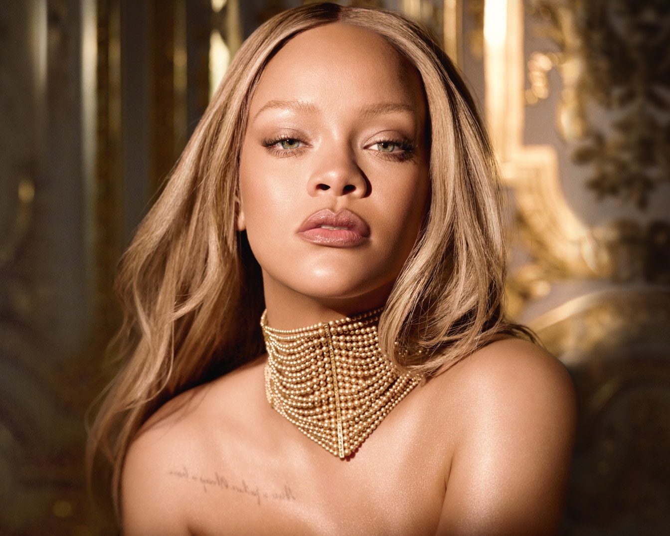 Close-up van Rihanna met losse blonde golven, een gouden kralenketting en een subtiele make-up look.