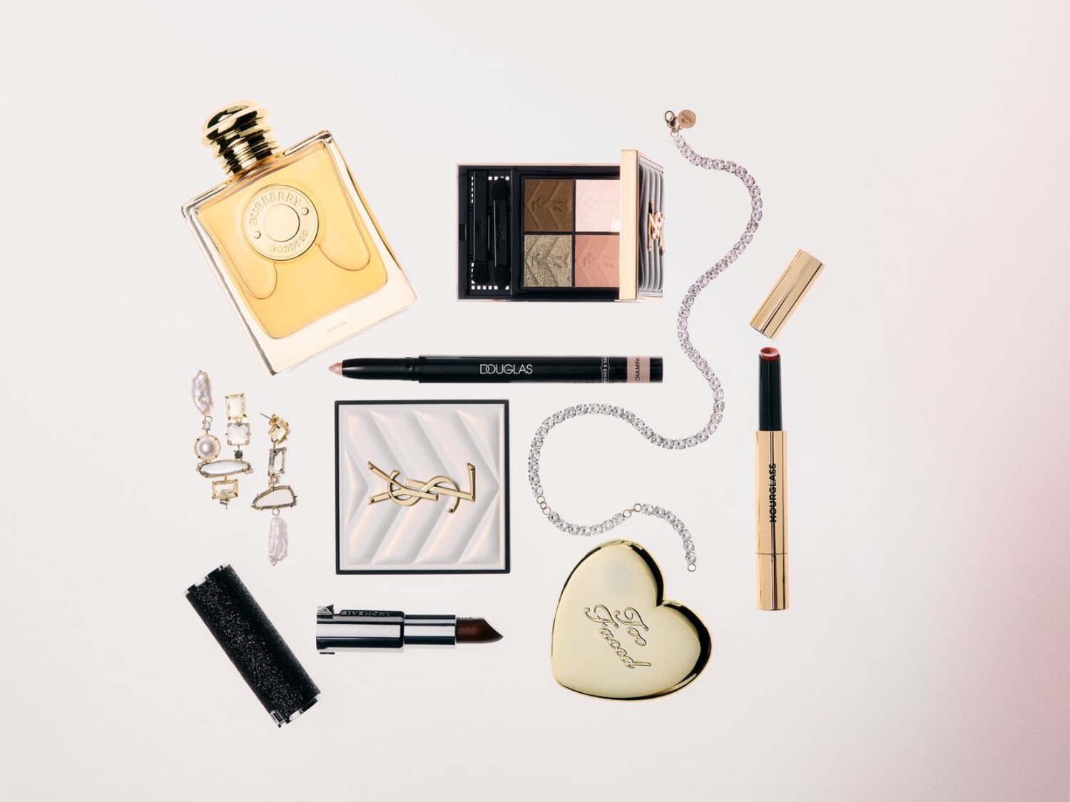 Elegancki zestaw kosmetyków i biżuterii, w tym perfumy Burberry Goddess w złotej butelce, paleta cieni do powiek, szminka Hourglass w złotym opakowaniu i błyszczący naszyjnik.