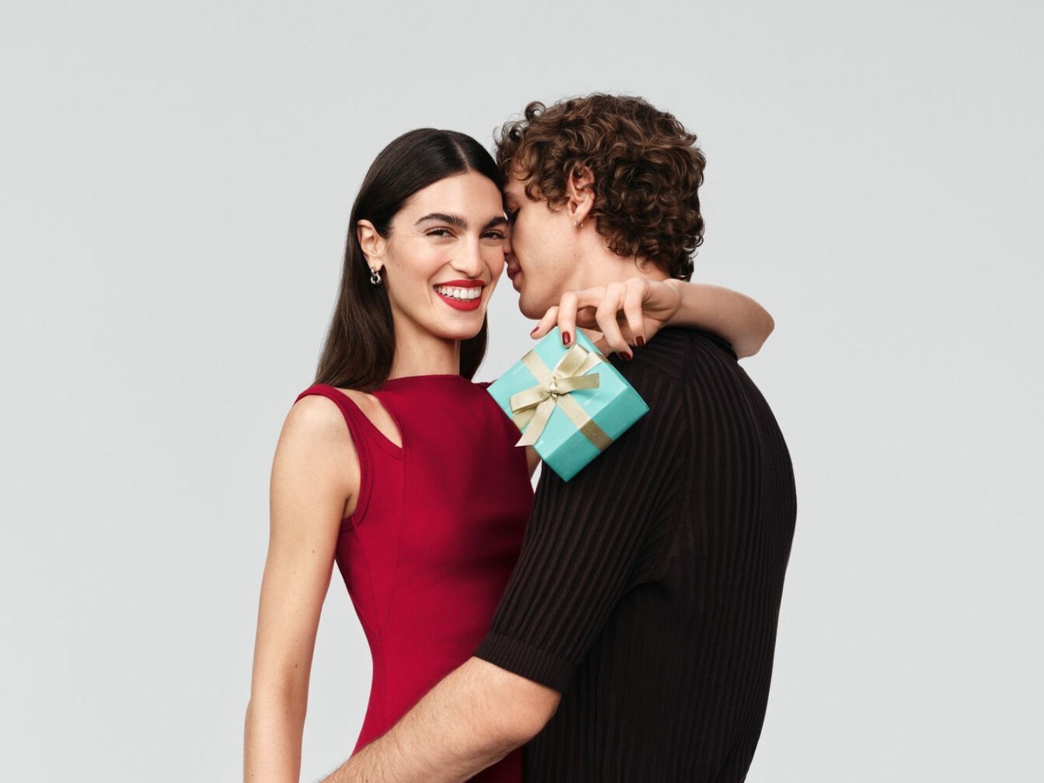 Uśmiechnięta kobieta w czerwonej sukience trzymająca turkusowe pudełko z kokardą, wtulona w mężczyznę w czarnym swetrze.