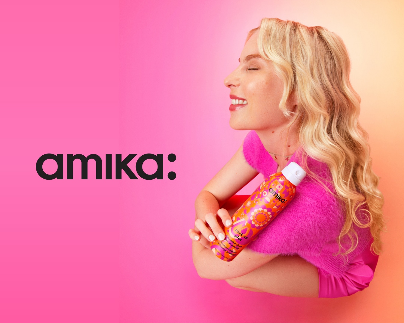 Une femme blonde souriante, les yeux fermés, serre contre elle un aérosol orange à motifs floraux de la marque Amika, sur un fond rose vif.