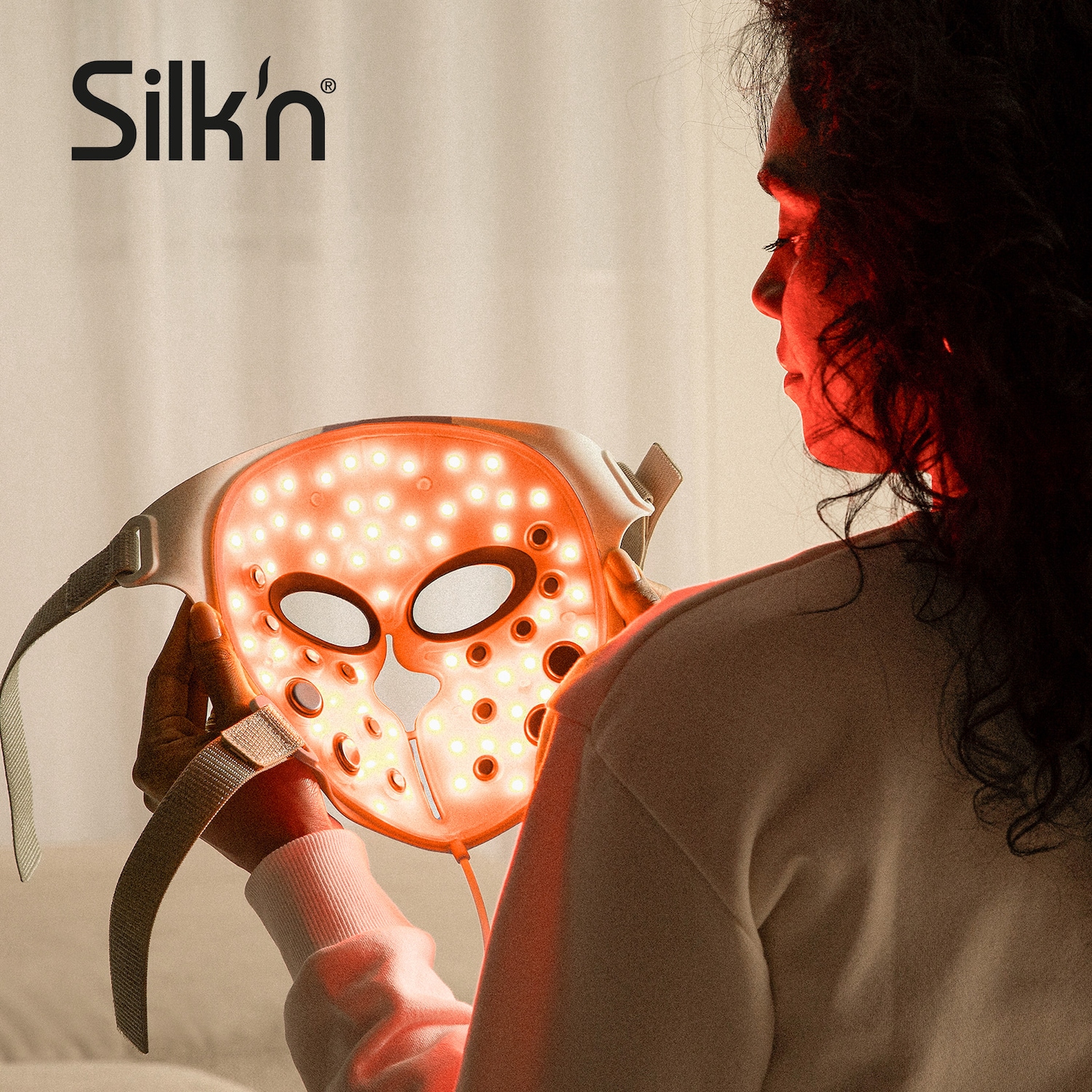 Une femme tient un masque de luminothérapie Silk'n illuminé d'une lumière rouge, promettant une peau éclatante et rajeunie.