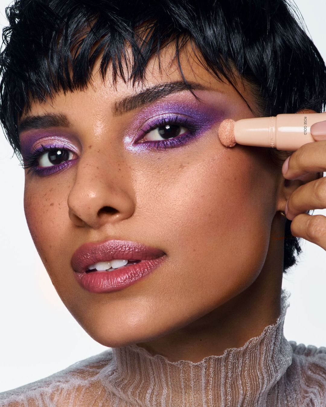 De cerca, una mujer aplica sombra de ojos rosa dorada con un aplicador en barra sobre un maquillaje de ojos morado brillante y labios rosados brillantes.