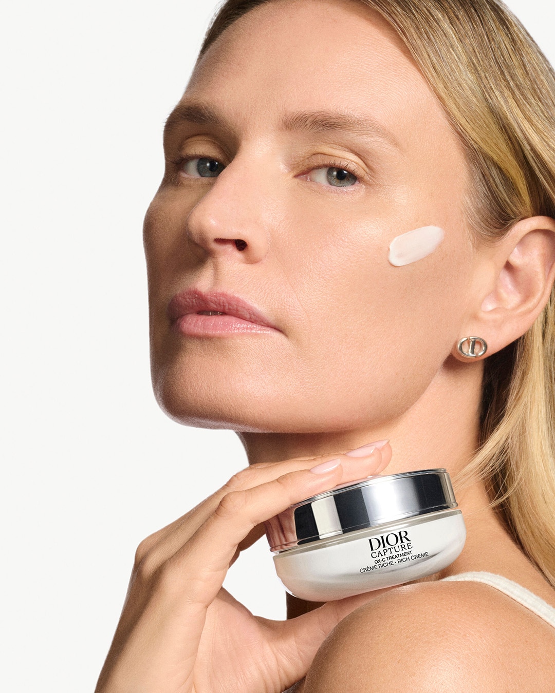 Una mujer rubia con un pendiente de Dior en la oreja derecha sostiene un tarro blanco de crema Dior Capture Totale con tapa plateada, mientras una pequeña cantidad de crema se aplica en su mejilla derecha.