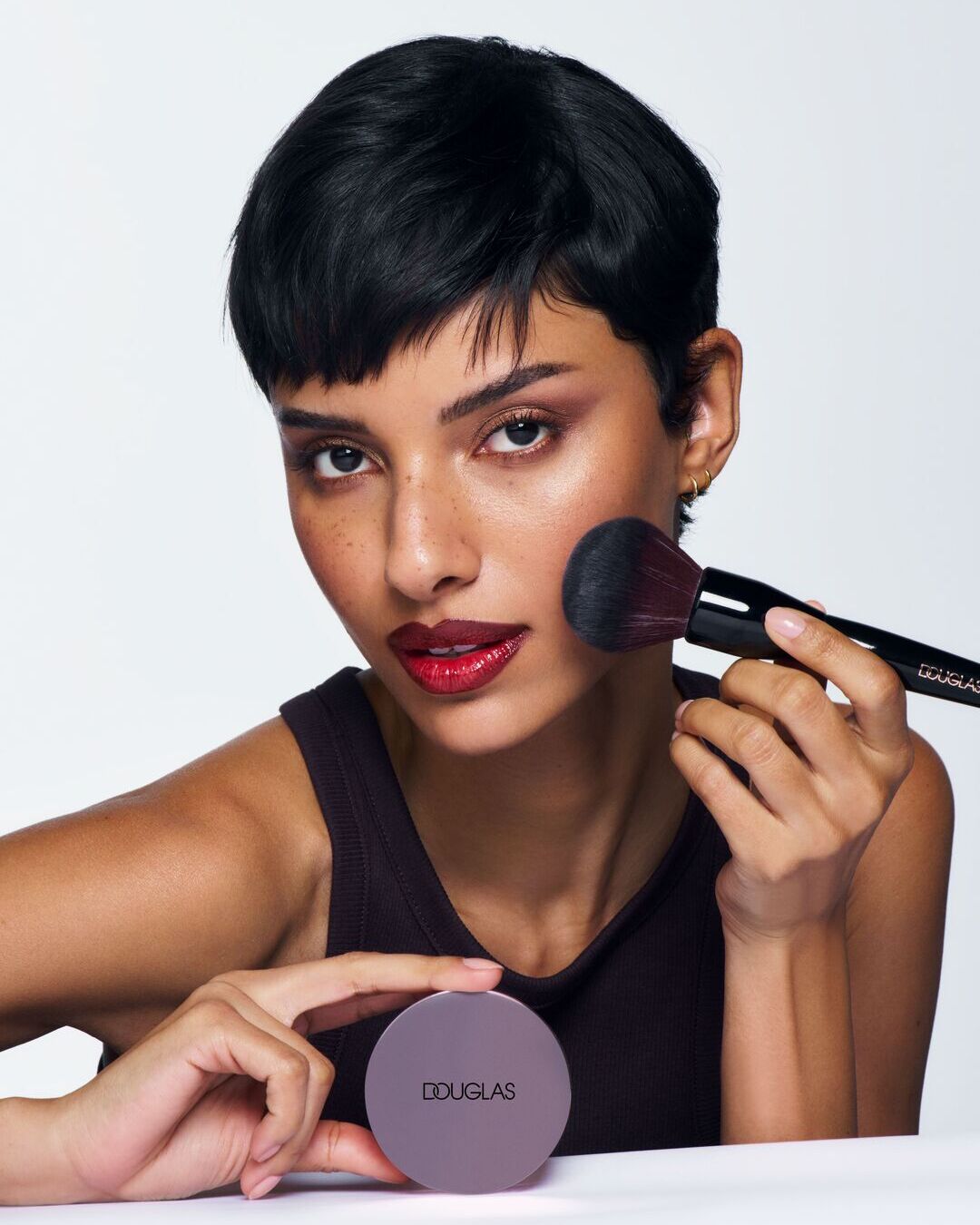 Una mujer de piel morena se aplica maquillaje con una brocha y sostiene un compacto de Douglas, resaltando su belleza con un toque de color.