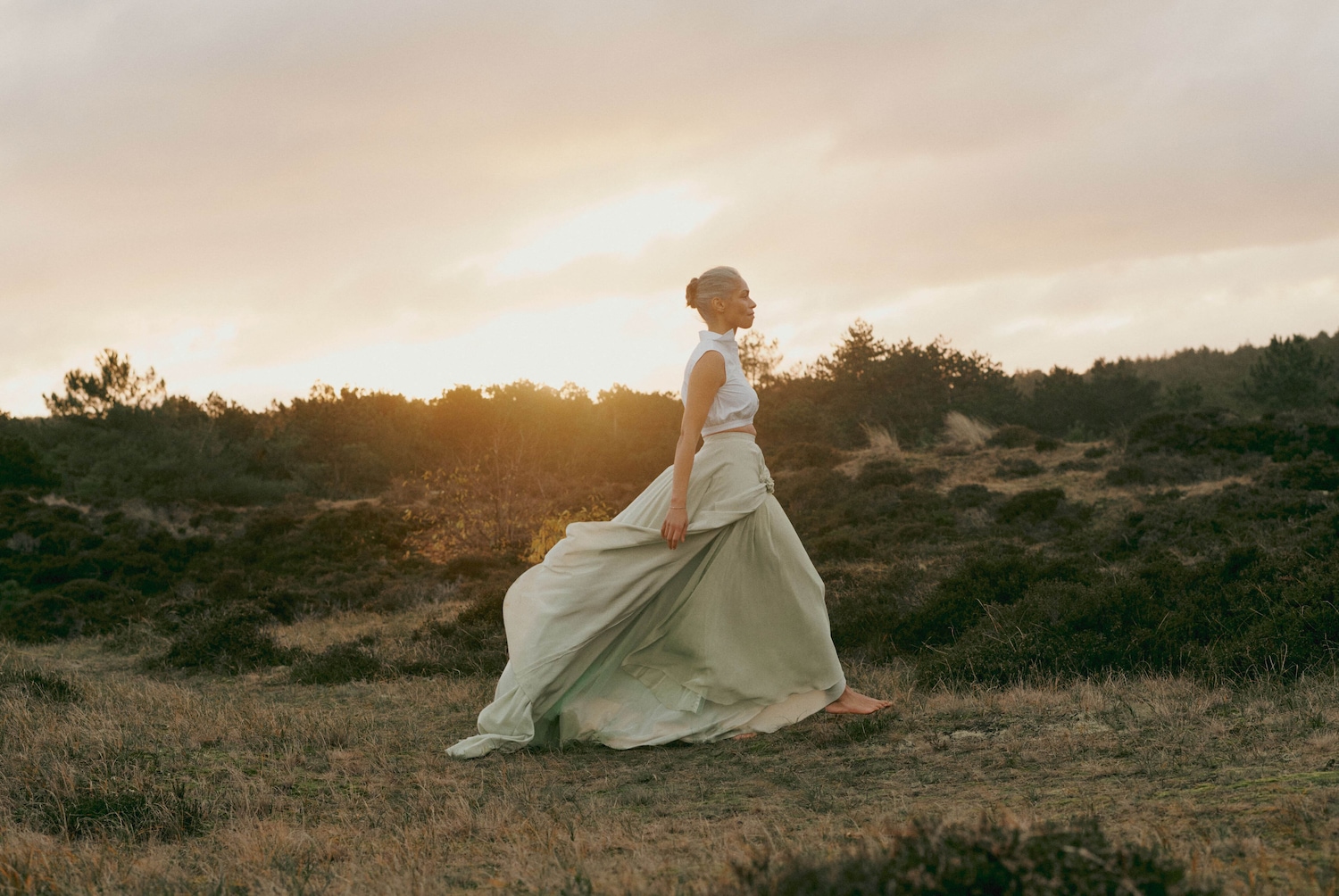 Eine Frau mit hochgesteckten grauen Haaren, bekleidet mit einem weissen Oberteil und einem fliessenden, hellgrünen Rock, geht barfuss durch eine natürliche Landschaft im sanften Licht der Abenddämmerung.