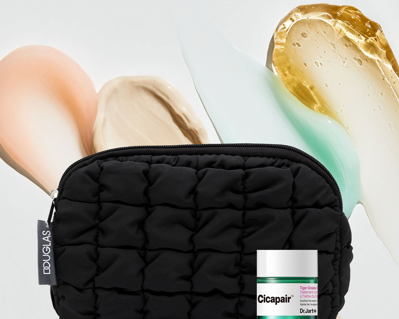 Eine schwarze Kosmetiktasche von Douglas liegt vor einem Hintergrund mit verschiedenen Cremetupfern in Gelb-, Beige- und Rosatönen, zusammen mit einer grünen Cicapair Creme von Dr. Jart+.