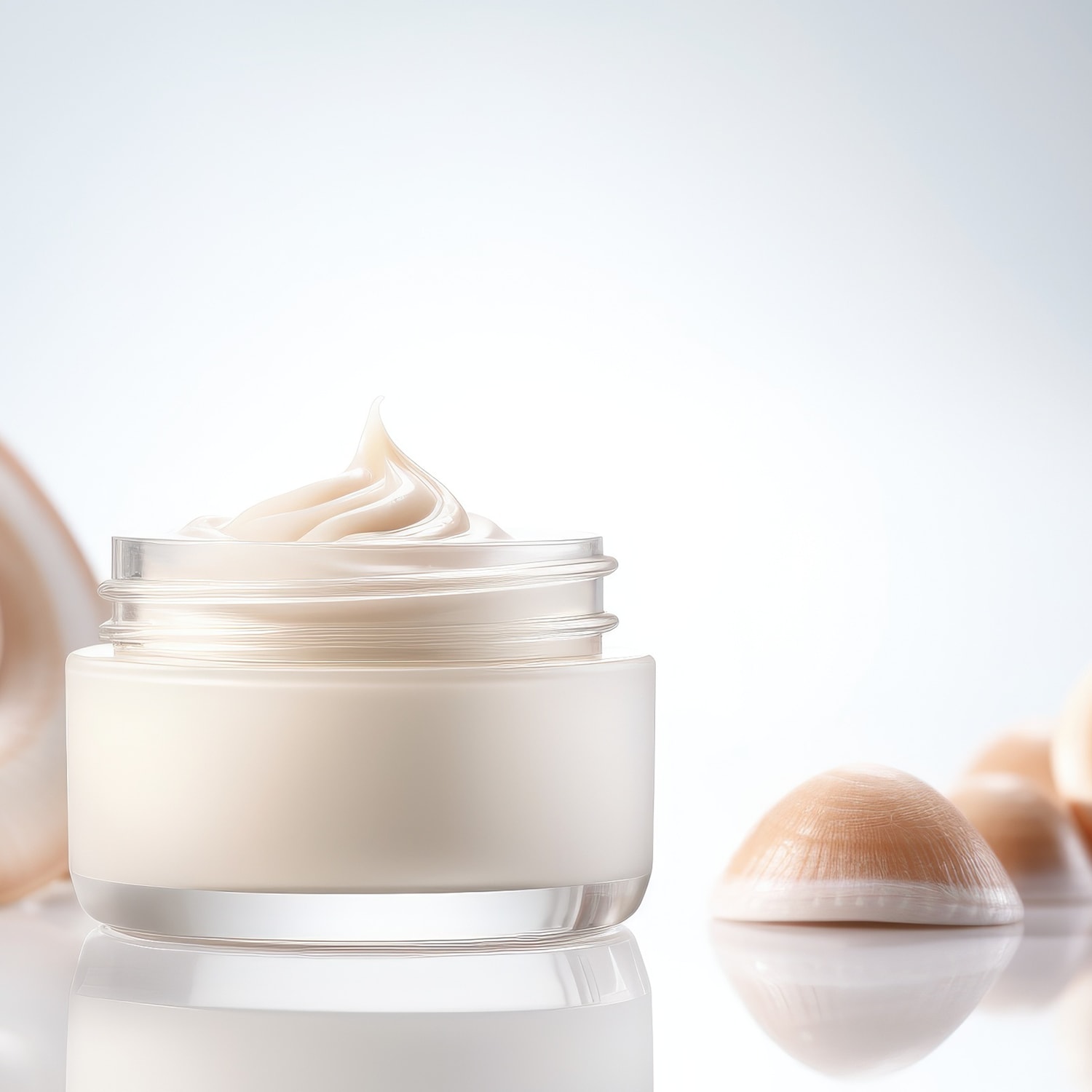 Nahaufnahme einer durchsichtigen Glasdose mit beiger Creme, die spiralförmig aufgetragen ist, umgeben von Muscheln auf einer glänzenden Oberfläche.