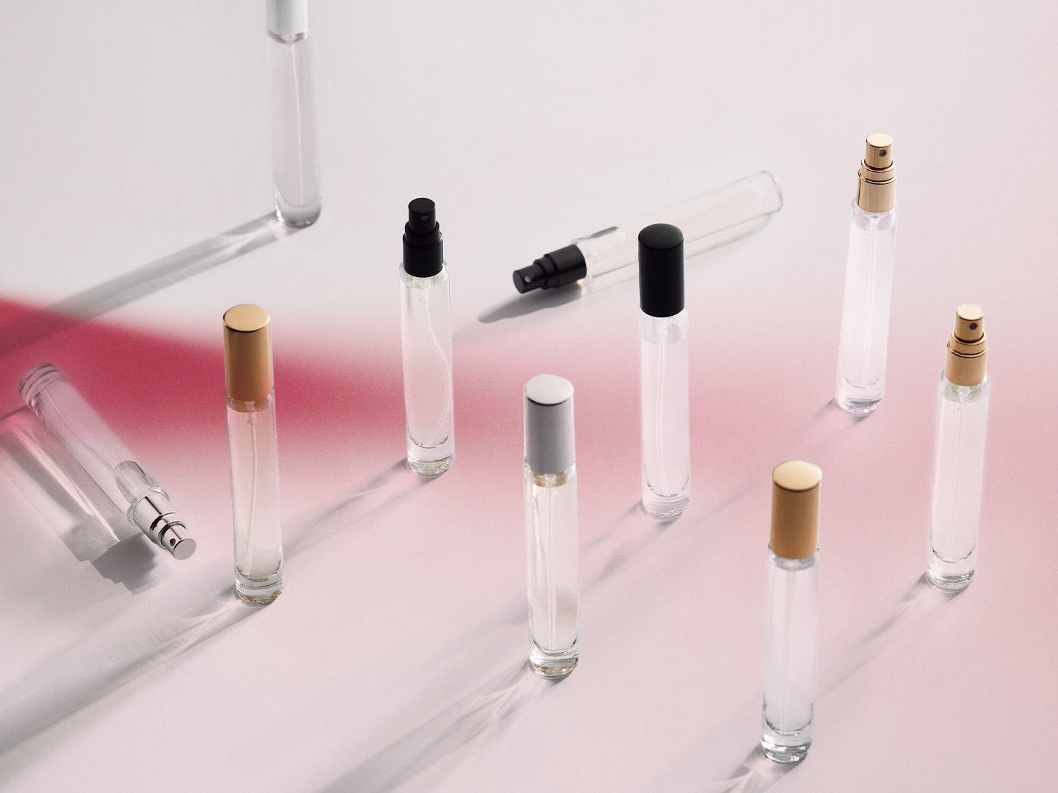 Verschiedene durchsichtige Parfümflaschen mit silbernen, goldenen und schwarzen Kappen stehen auf einer hellrosa Oberfläche.