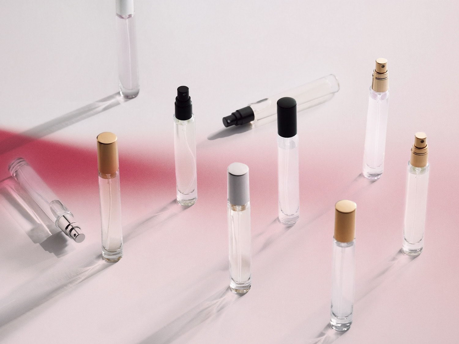 Verschiedene durchsichtige Parfümflaschen mit silbernen, goldenen und schwarzen Kappen stehen auf einer hellrosa Oberfläche.
