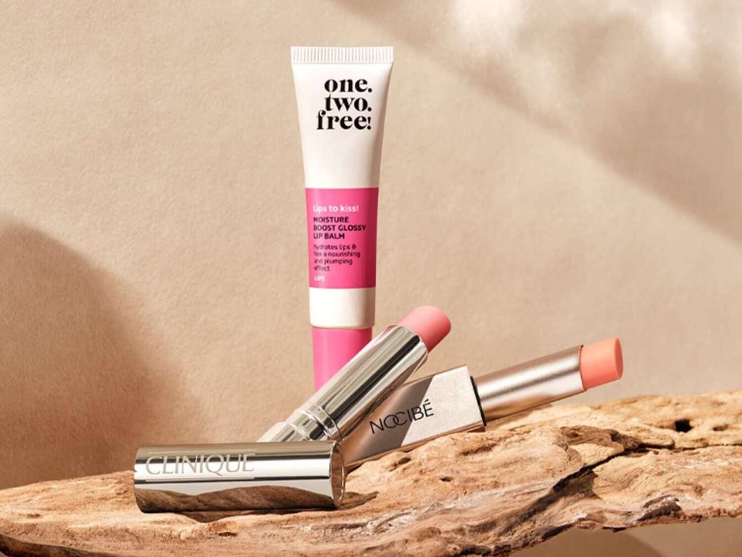 Découvrez le baume à lèvres hydratant One.Two.Free! dans son tube blanc et rose, ainsi que les rouges à lèvres Clinique et Nocibé aux teintes délicates, présentés sur un morceau de bois naturel.