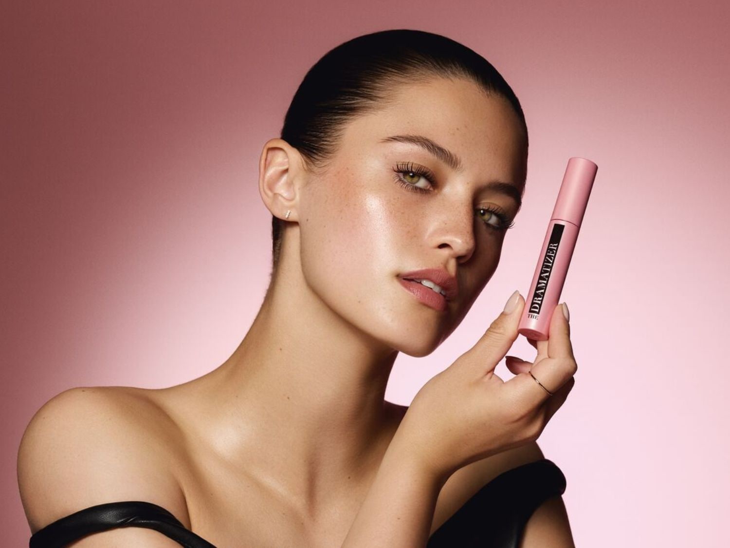 Une femme tient un mascara rose Dramatizer, avec un fond rose doux qui met en valeur le produit et son regard captivant.