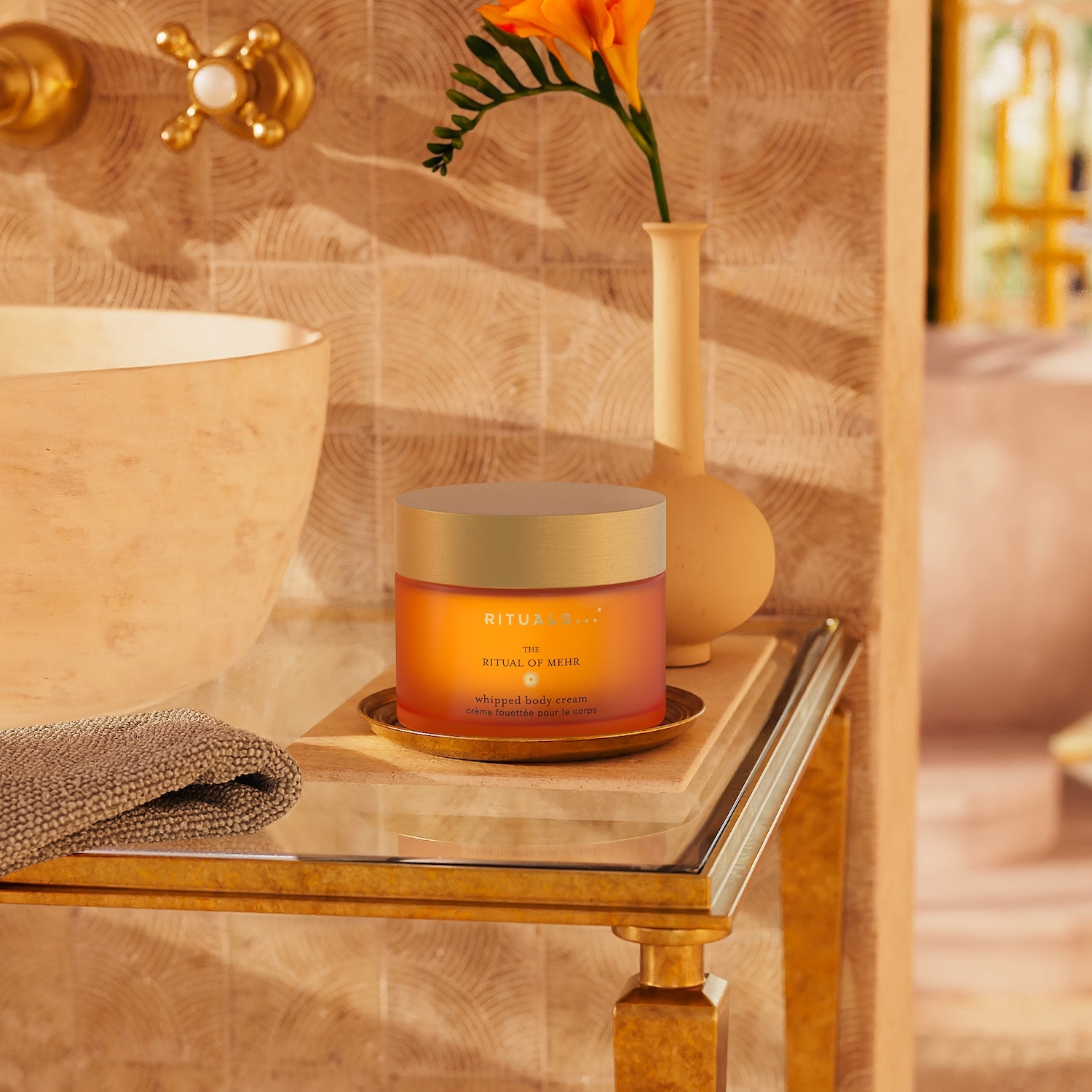 Ein Tiegel der Ritual of Mehr Whipped Body Cream steht auf einem goldenen Tisch neben einem beigen Waschbecken und einer Vase mit einer orangefarbenen Blume.