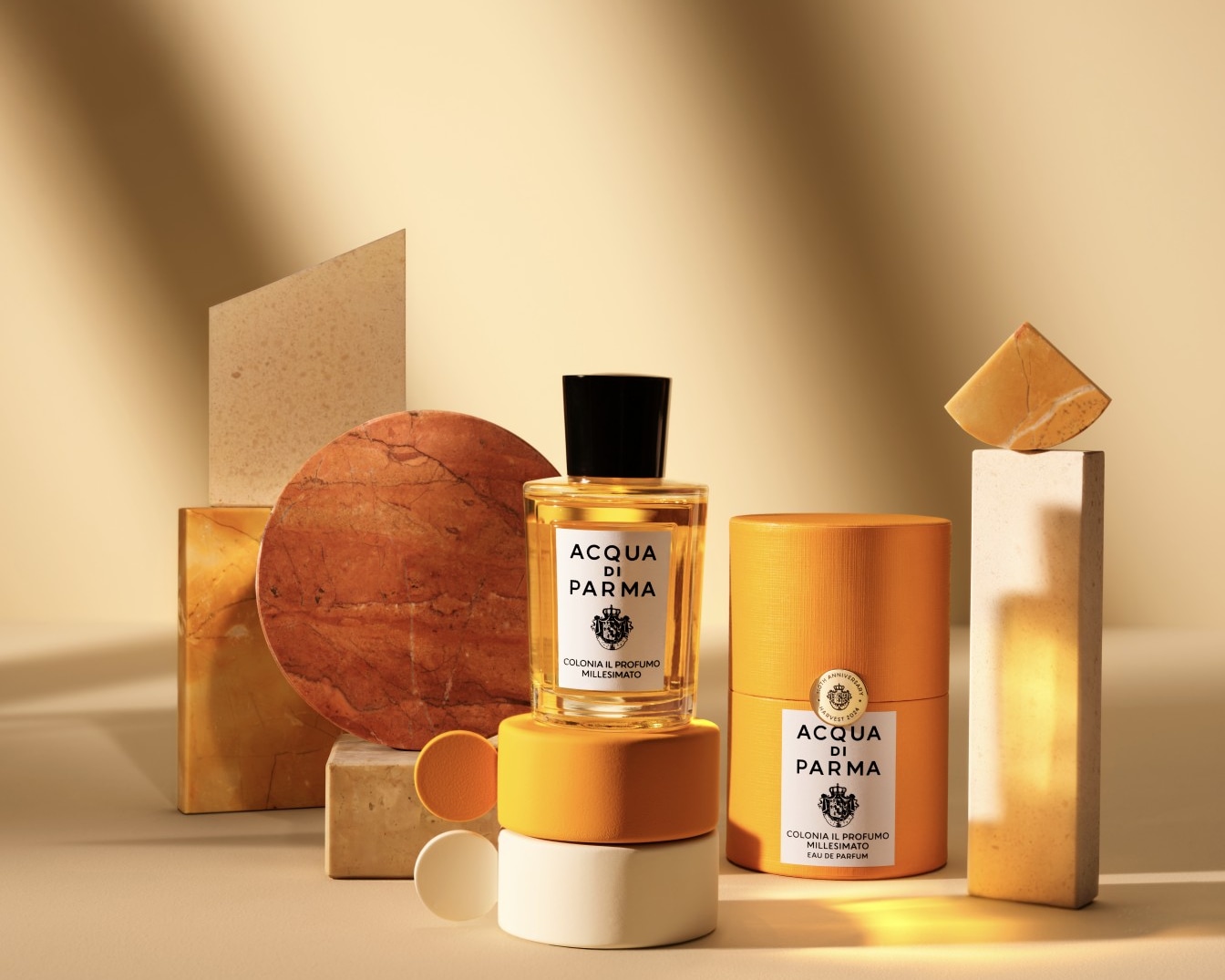 Flakon wody kolońskiej Acqua di Parma Colonia Il Profumo Millesimato z czarną zakrętką i pomarańczową etykietą, stoi na pomarańczowym pudełku na tle beżowych i pomarańczowych elementów dekoracyjnych.