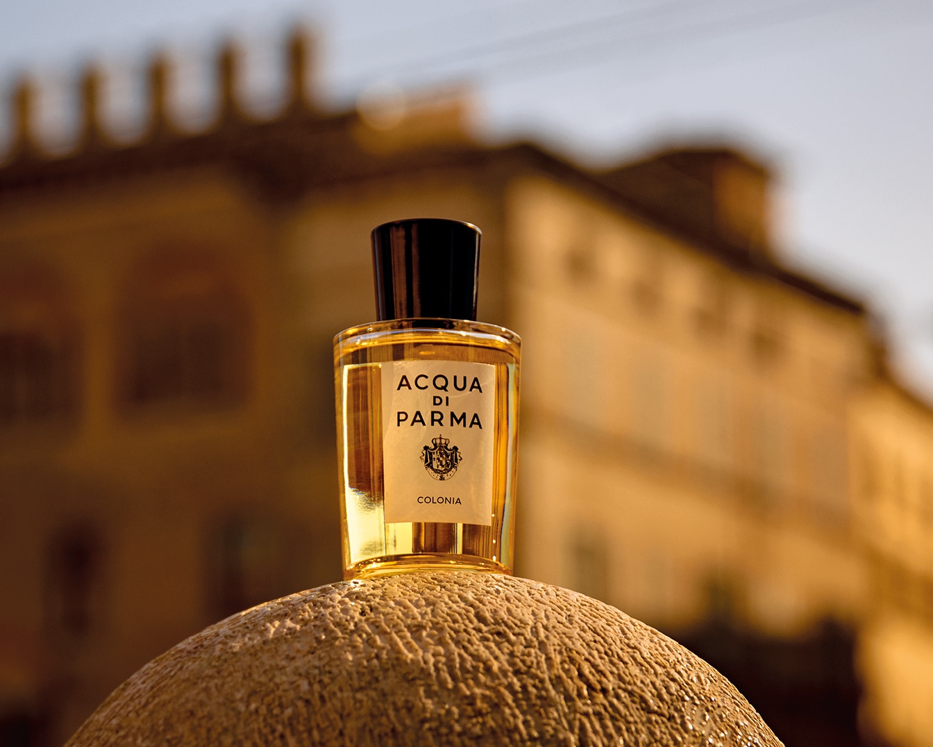 Butelka wody kolońskiej Acqua di Parma stoi na kamiennej powierzchni, z rozmytym budynkiem w tle, co dodaje jej elegancji.