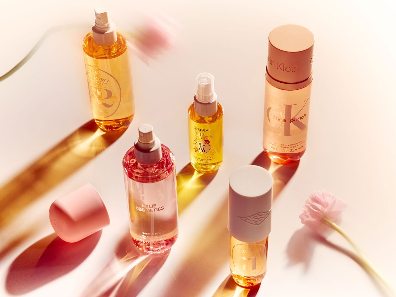 The image displays a collection of body mists and perfumes from various brands like Sol de Janeiro, Kylie Cosmetics Vanilla Dew, Douglas, and Calvin Klein Sheer Peach, each in uniquely designed bottles.